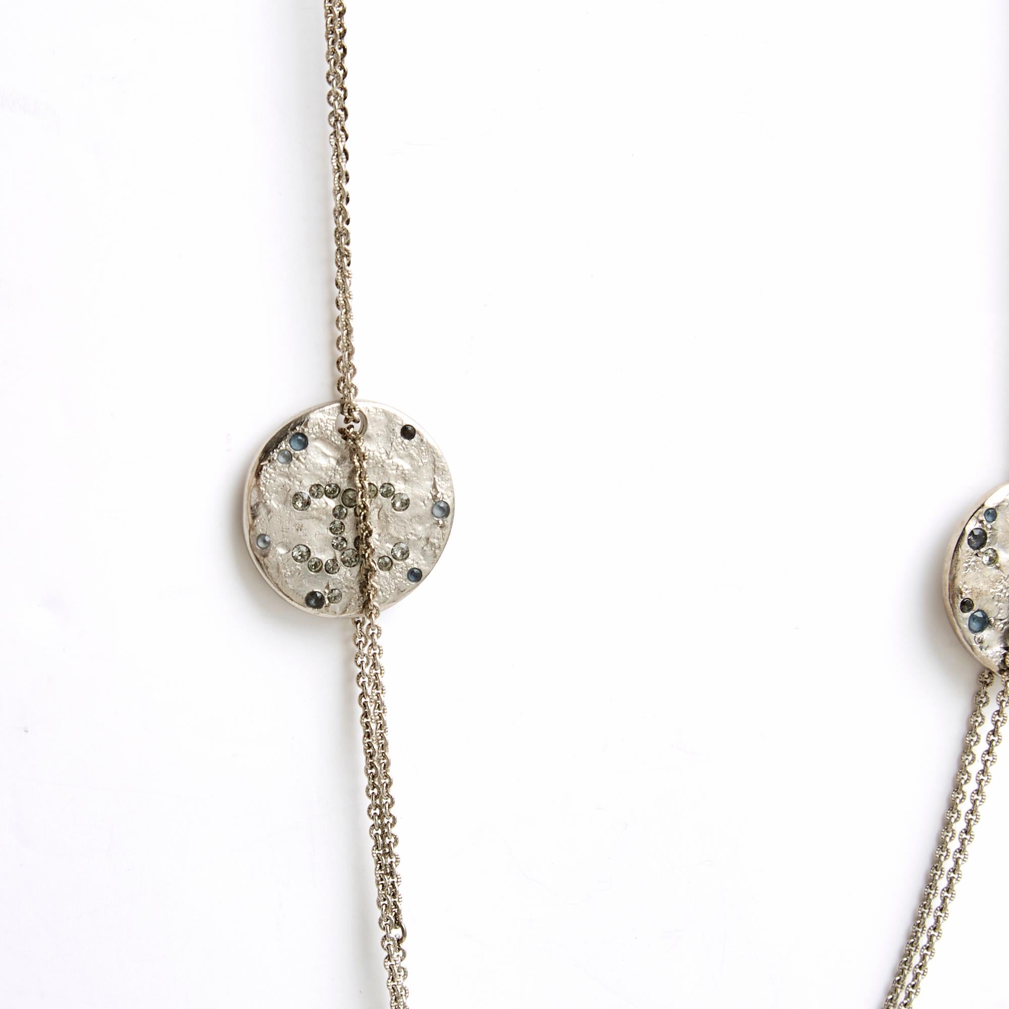 Collana sautoir Chanel della collezione SS2016 in metallo argentato composta da 2 catene con anelli lavorati e decorati con medaglioni o pezzi del logo CC incrostati di strass grigi di diverse dimensioni e forme, che si chiudono con un moschettone