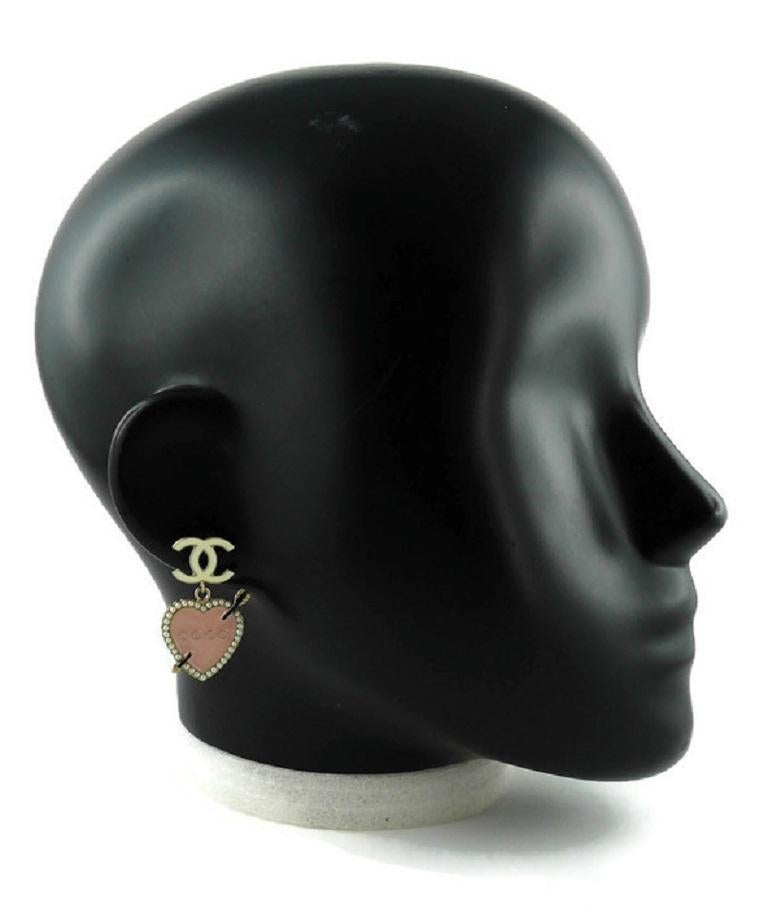 CHANEL pale gold toned COCO dangling earrings (for pierced ears) featuring baby pink enamel heart adorned with faux pearls and shooting arrow, white CC logo top.

From the Spring 2010 Collection.

Marked CHANEL 10P Made in Italy.

Indicative