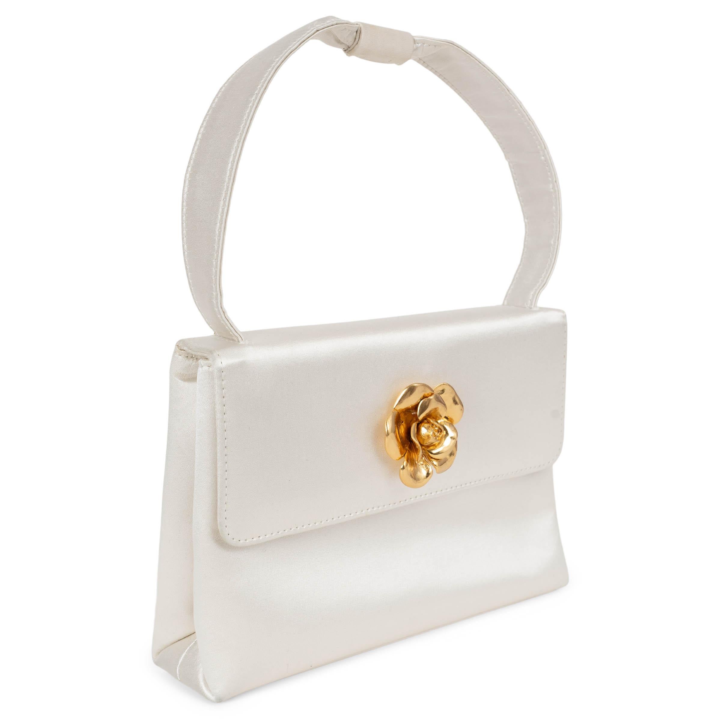 Este bolso de noche Chanel 100% auténtico está confeccionado en lustroso satén blanco nacarado y acentuado con una llamativa Camelia de metal dorado en el centro de la solapa. Un asa plana y un bolsillo trasero exterior añaden practicidad a su