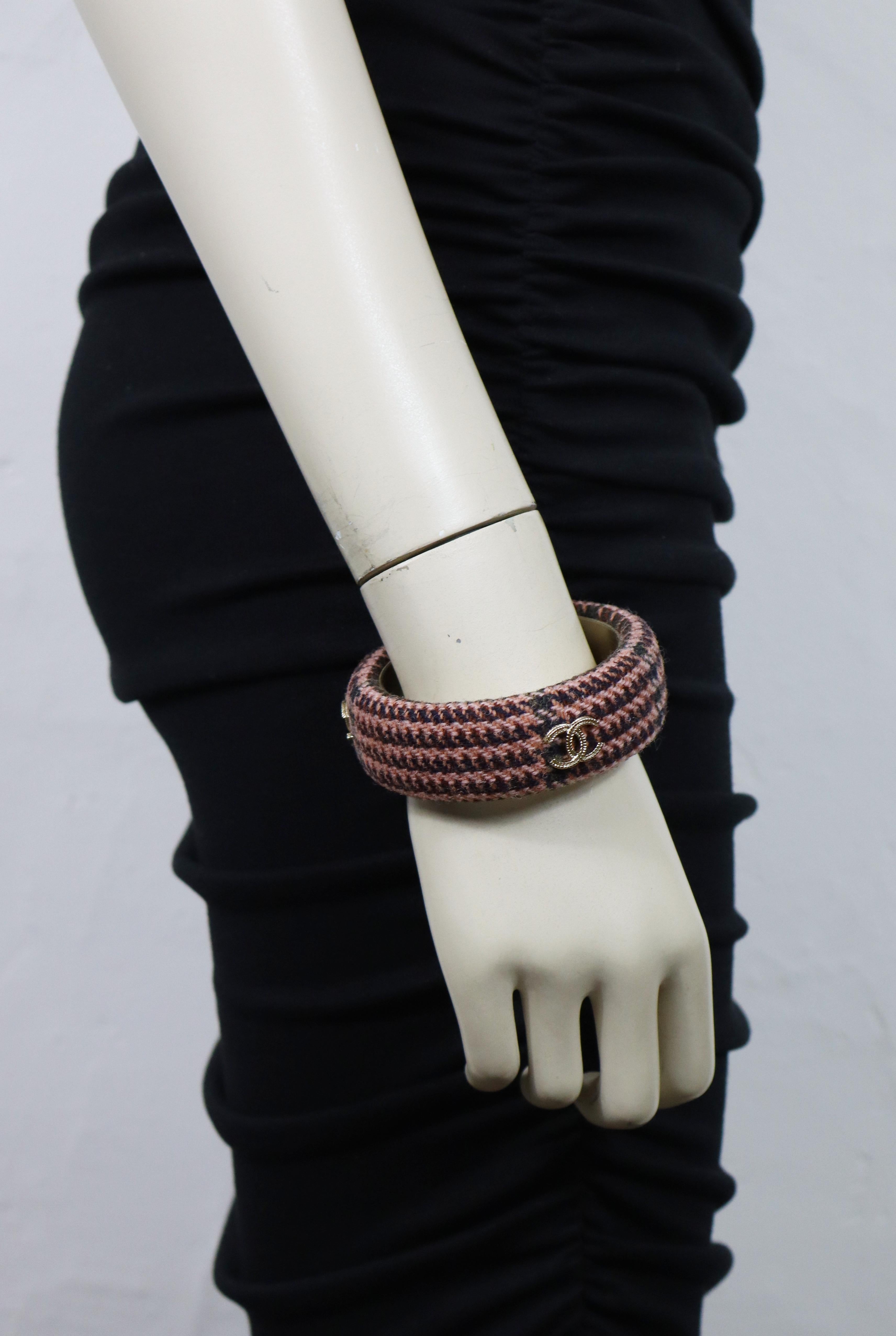 Bracciale bangle di Chanel in tweed di lana intrecciata nei toni del rosa e del marrone, con tre loghi CC dorati.

Marchio laser Chanel B13 A Made in Italy.
Il marchio della taglia recita : XS.
Marchio S di vendita privata inciso sul lato interno