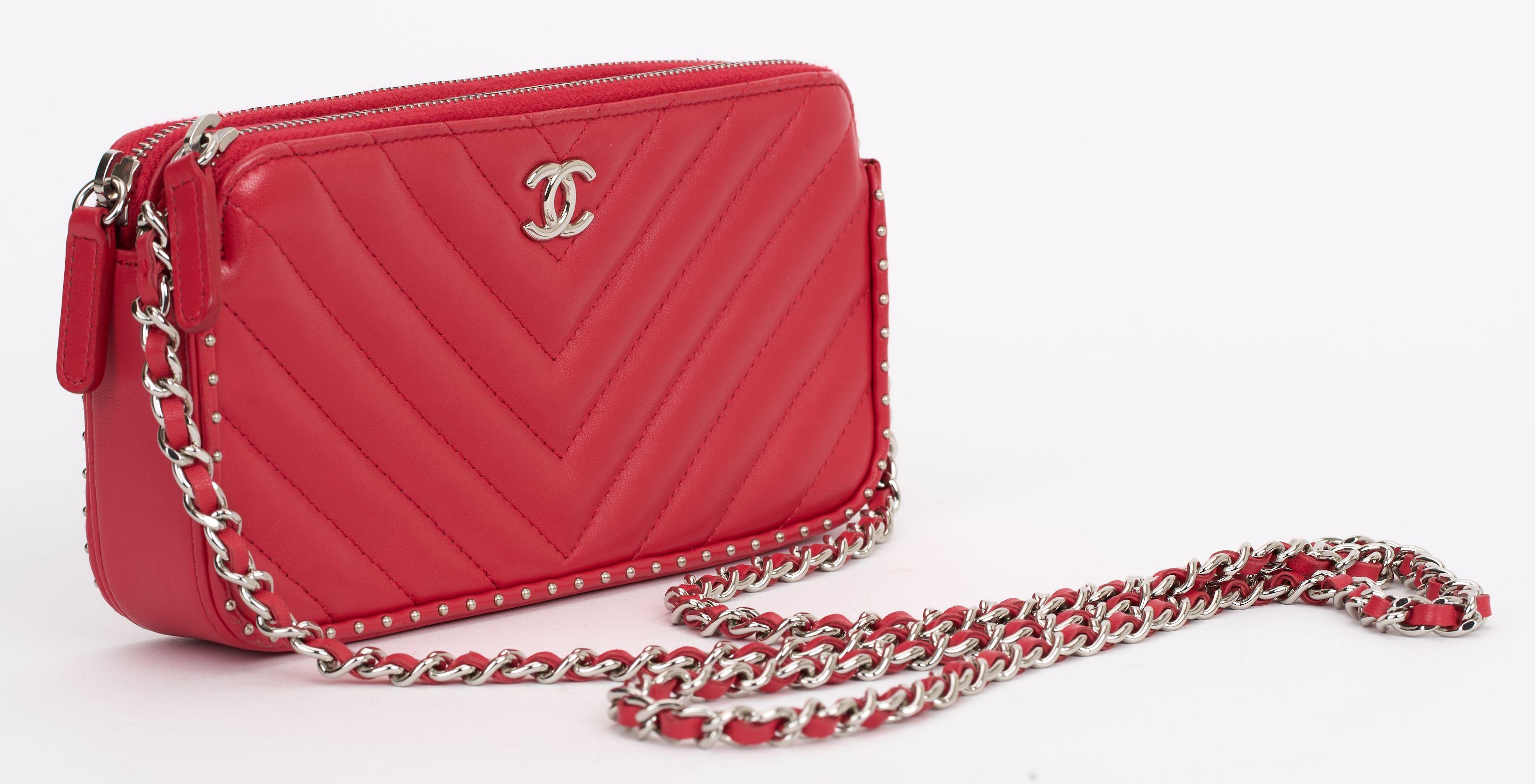 Este bolso cruzado Chanel Chevron de piel de cordero roja con tachuelas tiene una cadena extraíble y detalles de tachuelas en los bordes, con cierre de cremallera en la parte superior y seis ranuras para tarjetas en el interior. Este bolso cruzado