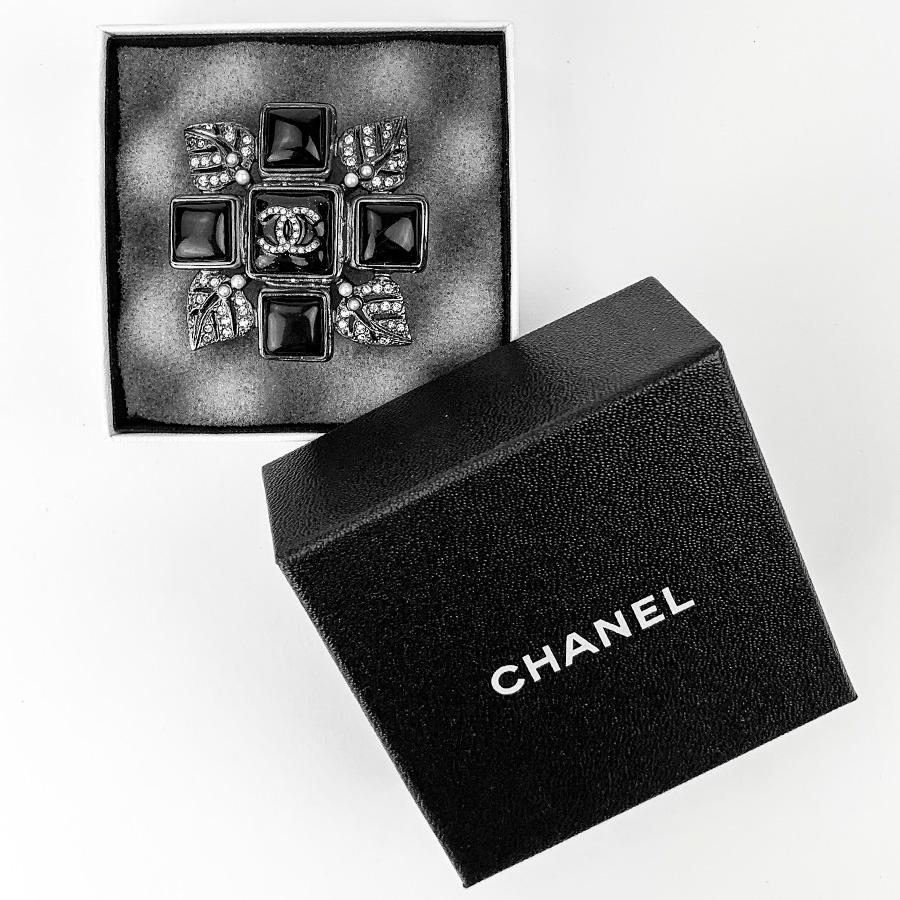 The brooch is signed by Maison CHANEL. It consists on a black resin cross around which come ruthenium metal sheets, set with rhinestones and pearls. We find the CC of the brand all in rhinestones in the center of the brooch.
The brooch is in very