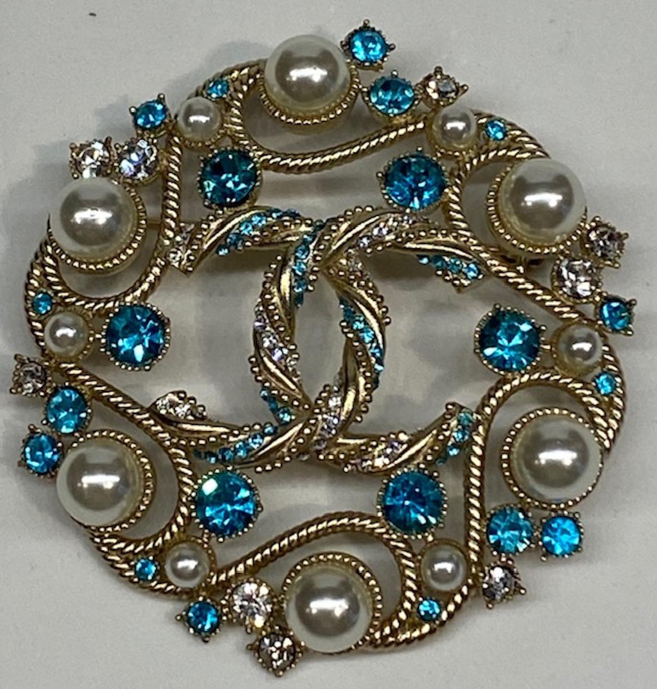A very very lovely Chanel round brooch in light gold and set with pearl, clear and aqua blue rhinestones from the 2019 Cruise Collection. Center of the pin features the iconic Chanel interlocking CC logo in twisted metalwork and set with tiny clear