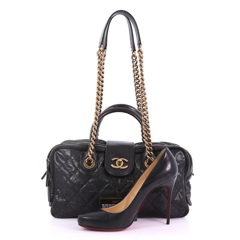 This Chanel Shiva Bowler Bag Quilted Caviar, crafted in black quilted caviar leather, features a chain-link shoulder strap with leather pads, dual rolled leather handles, leather tab with CC turn-lock closure, front zip compartment, and aged