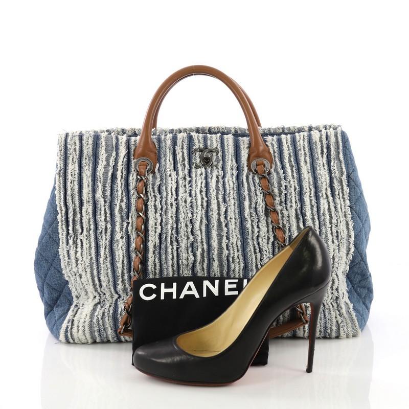 This Chanel Shopping Tote Fringe Denim Large, crafted in blue fringe denim, features dual rolled leather handles with woven-in leather chain straps, fringe denim details, side snap buttons, and aged silver-tone hardware. Its CC turn-lock closure