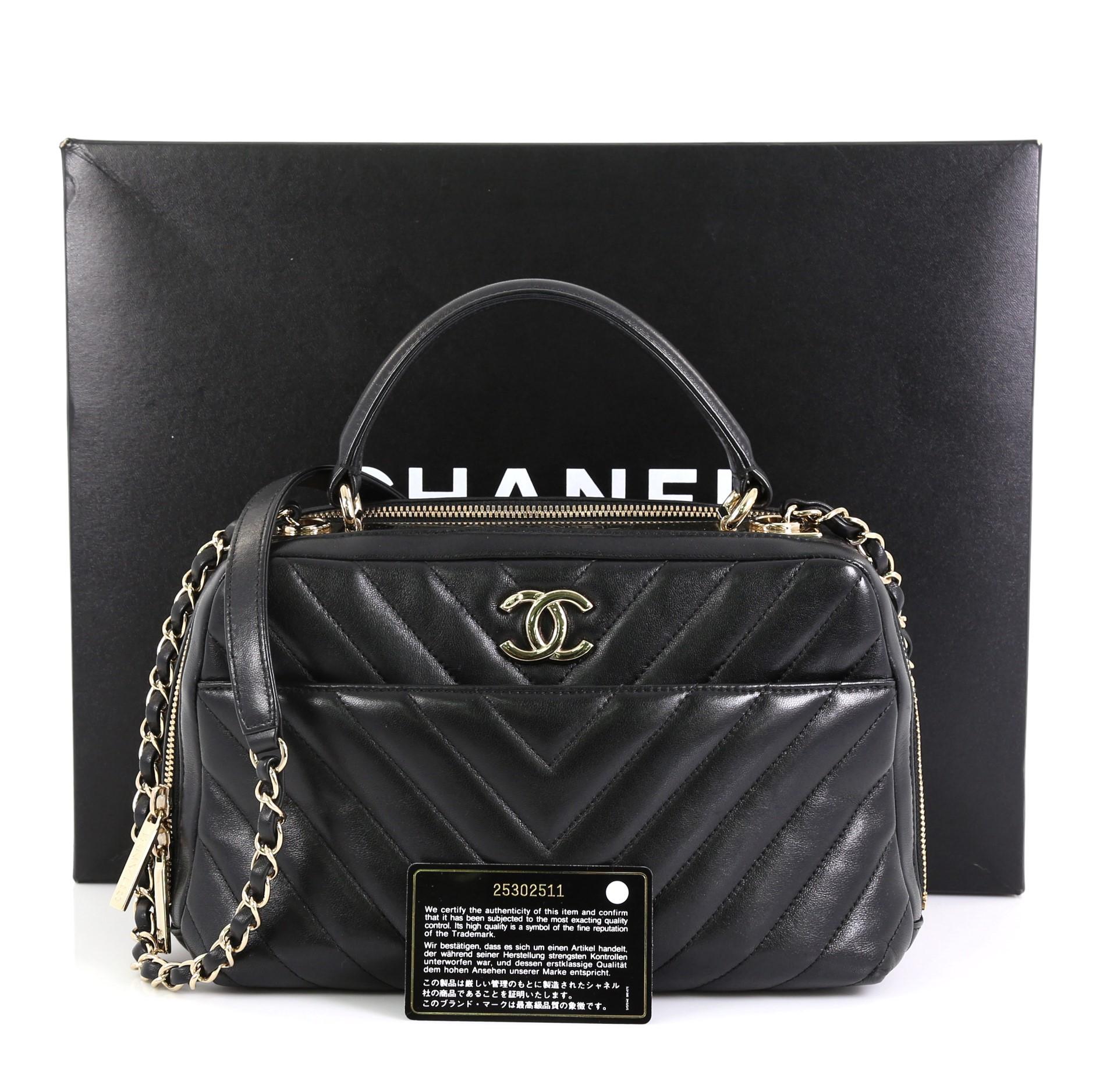 This Chanel Trendy CC Bowling Bag Chevron Lambskin Medium, crafted from black chevron lambskin leather, features a leather top handle, woven-in leather chain strap with leather pad, metal plate on top with Chanel logo, and gold-tone hardware. Its