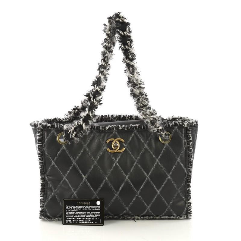 This Chanel Tweedy Tote Quilted Leather Medium, crafted from black quilted leather, features chain straps wrapped in tweed, tweed fringe trim, protective base studs, and aged silver and gold-tone hardware. Its CC turn-lock closure opens to a black