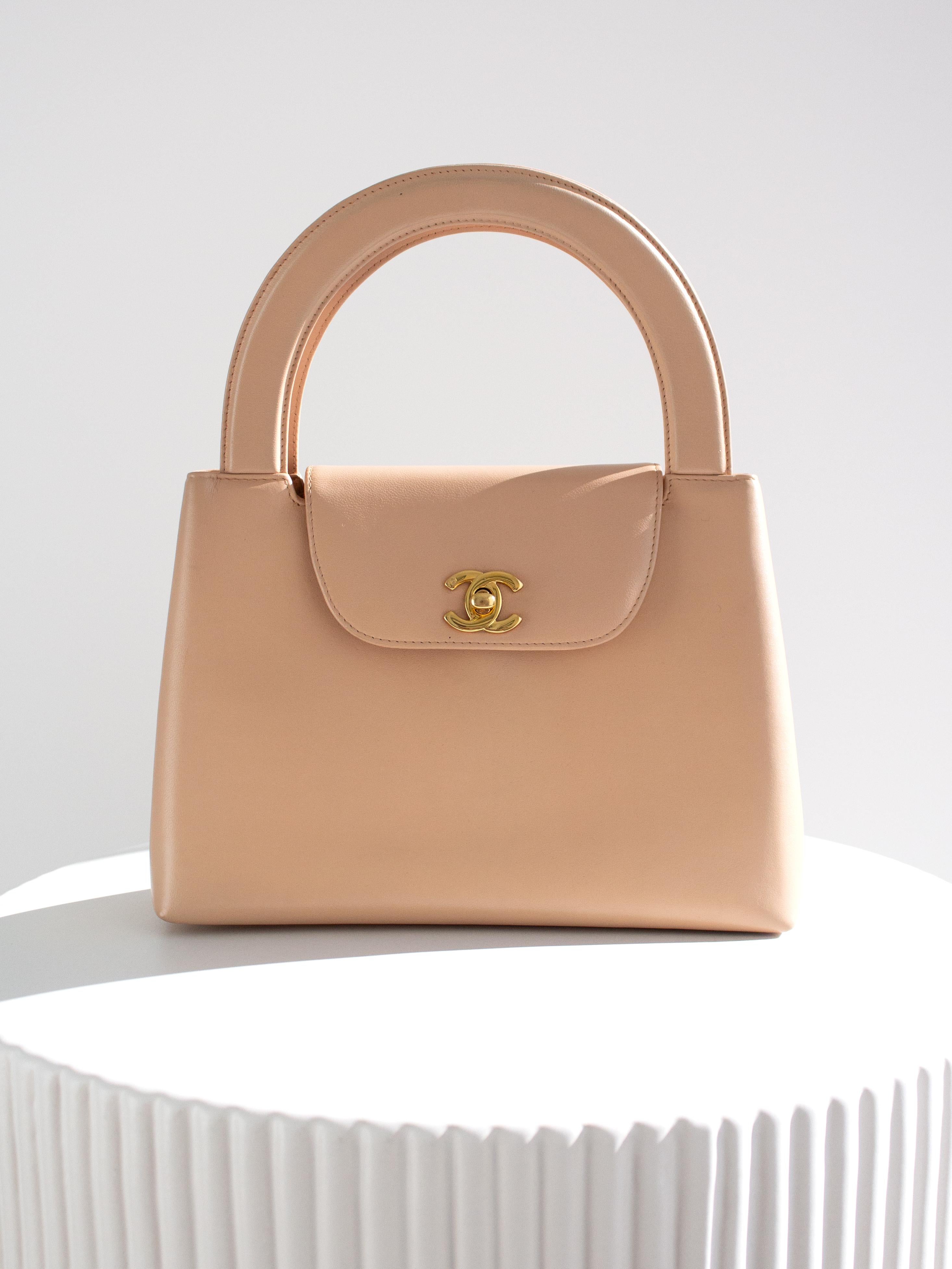 This 1998 Chanel Kelly top handle bag is a rare gem in a soft nude pink hue, a delicate blend of pink with a hint of beige. Crafted from buttery soft lambskin leather, the bag is adorned with elegant 24K gold-plated hardware, including the iconic CC