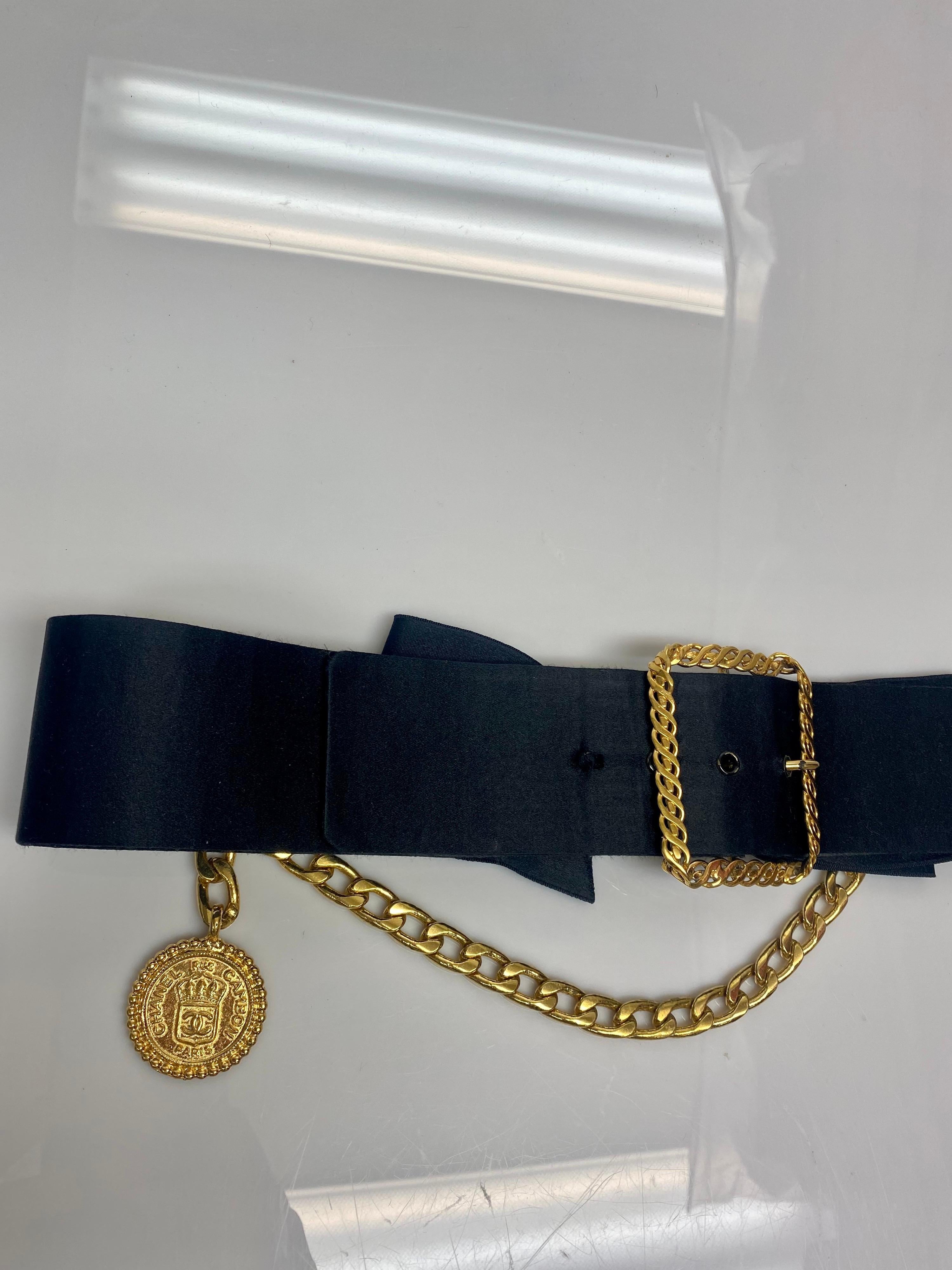 Ceinture Vintage By By en satin noir avec boucle dorée, chaîne et médaillon. Cette ceinture vintage de Chanel est un ajout luxueux à toute garde-robe. L'article comporte un nœud en satin noir et une grande boucle en métal doré avec une chaîne et un