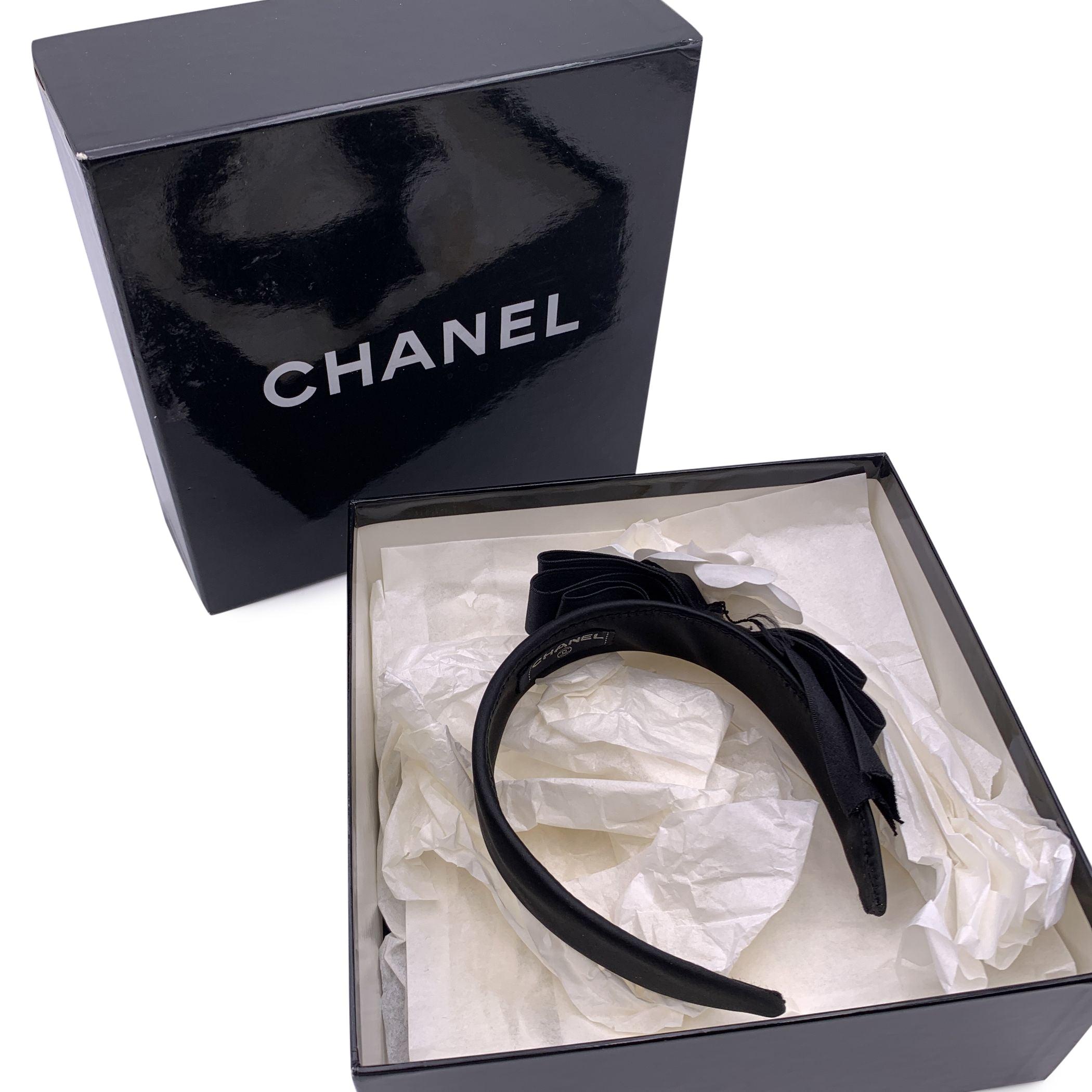 Lovely Chanel headband in black silk satin embellished with the a bow and a white camellia. 'Chanel' tag. Condition A - EXCELLENT In excellent condition. Chanel box included. Please check the photos carefully and ask if you need to know more details
