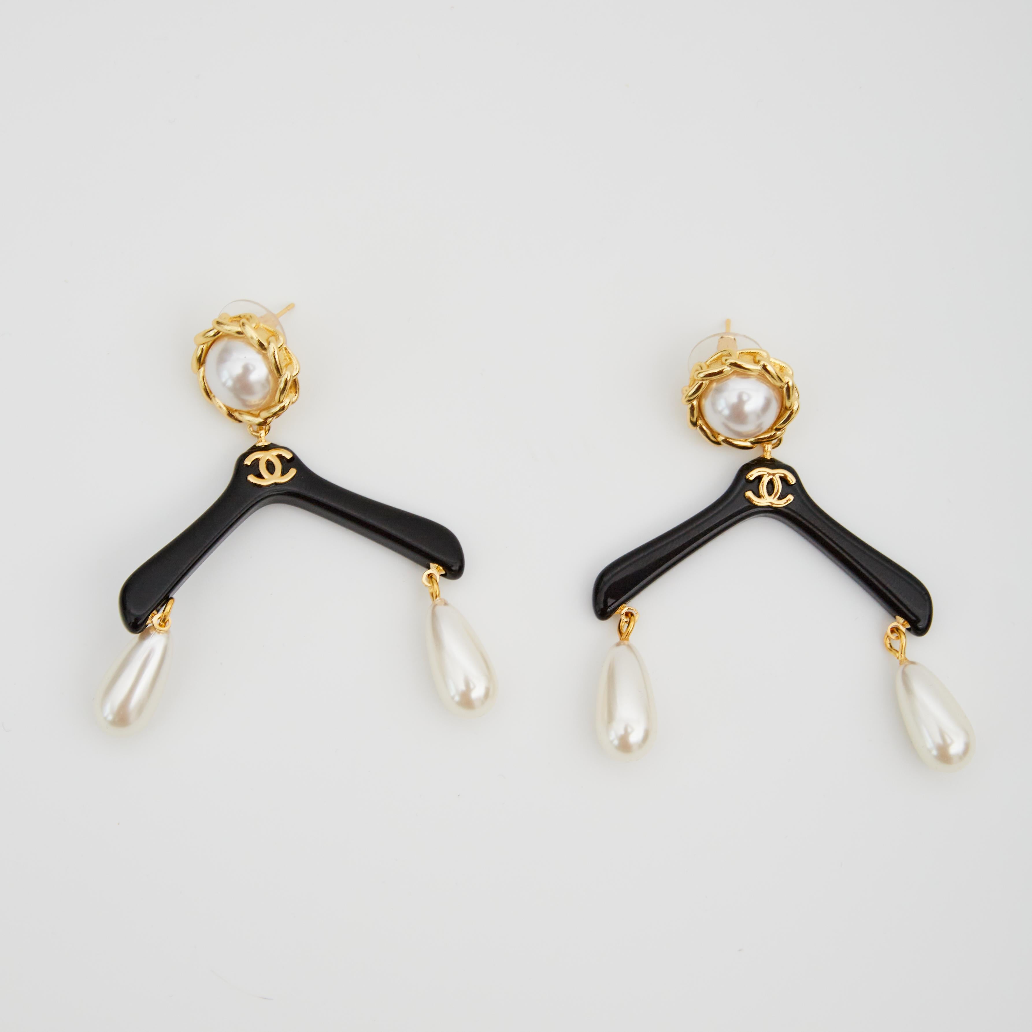 Chanel earrings fittings the shape of a coat hanged, pearl drops, CC logo and clip style attachment. 1980s

COLOR: Black and Gold
MATERIAL: Resin, faux pearl and brass
MARKINGS: A21 V
MEASURES: L 2.25” x L 2”
DROP: 2.25