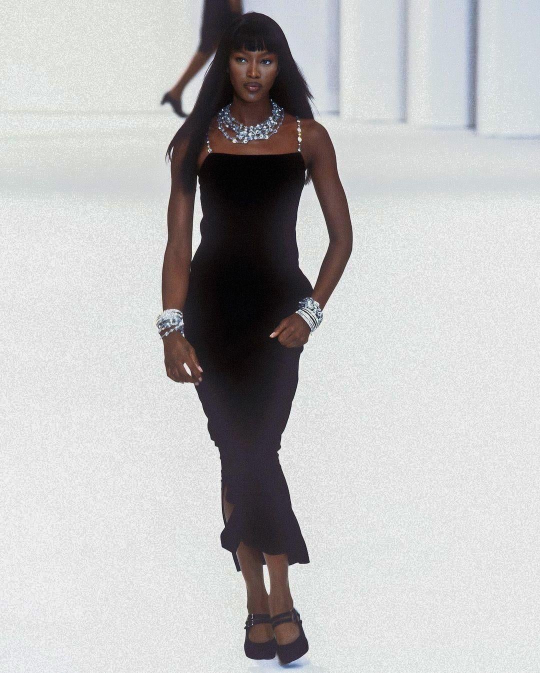 Meet this exquisite vintage Chanel dress from the iconic Spring/Summer 1998 collection, famously worn by the stunning Naomi Campbell. Crafted from luxurious black wool crepe, its silhouette is defined by silver tone chain straps adorned with