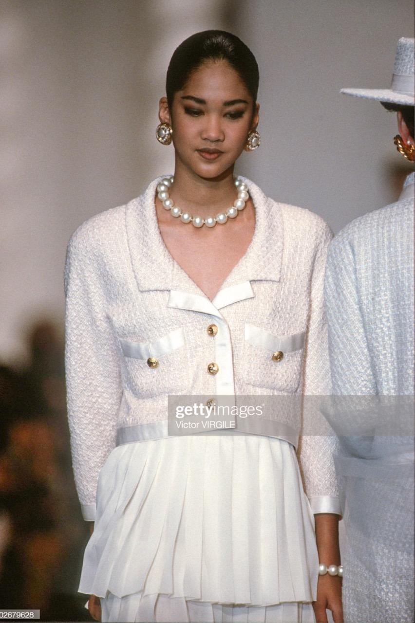 Iconic pink cropped jacket and mini skirt from Chanel Spring Summer 1990 collection. As seen on Claudia Schiffer. The suit is made of light pink and ecru tweed, adorned with white satin trims and signature gold CC buttons. Good vintage condition