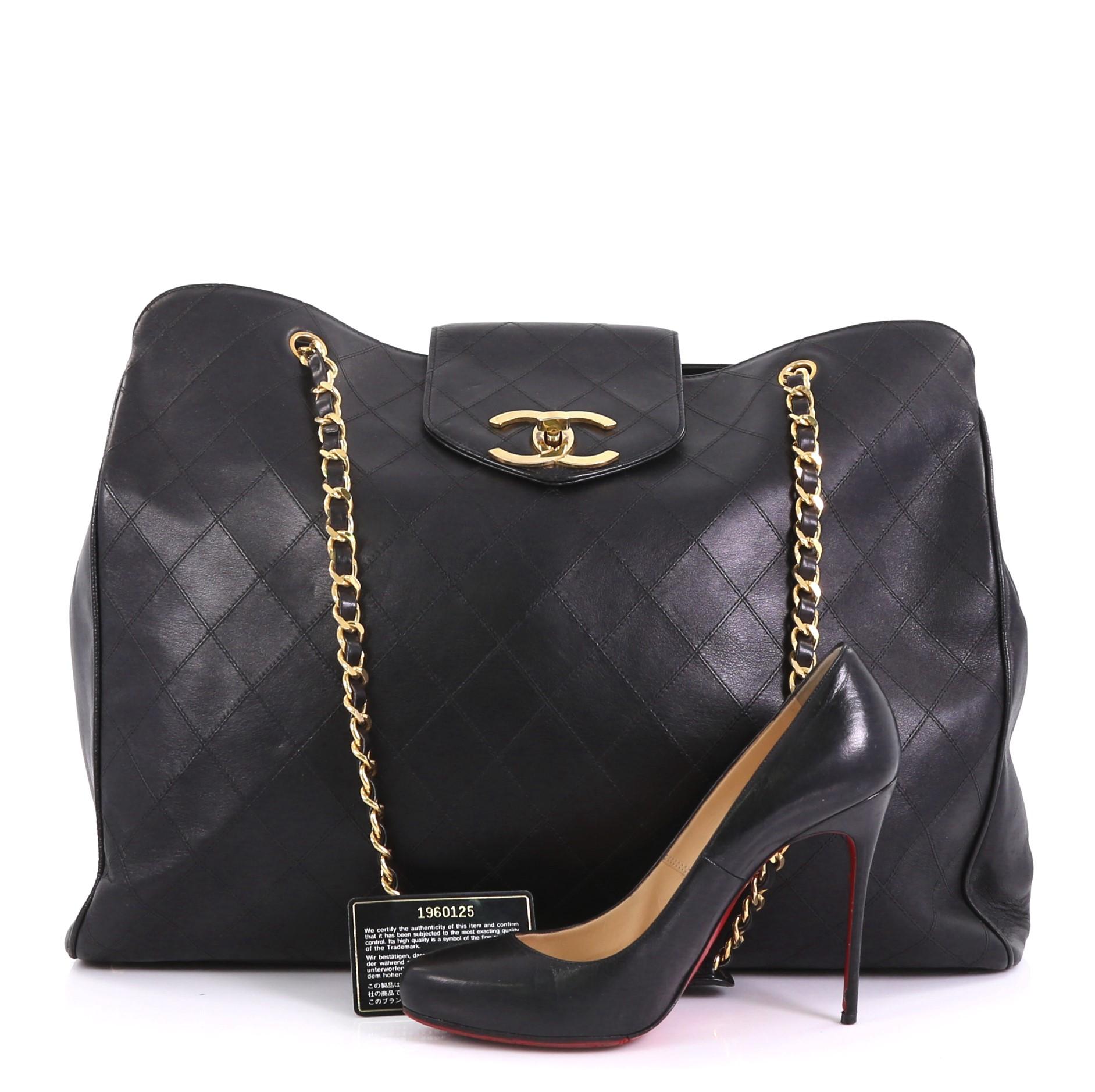 This Chanel Vintage Supermodel Weekender Bag Quilted Leather Large, crafted in black quilted leather, features dual woven-in leather chain straps, a flap tab with turn-lock closure, and gold-tone hardware. Its zip closure opens to a black leather