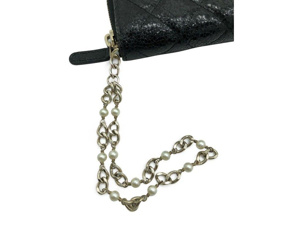 Este elegante bolso/cartera Chanel está confeccionado en lujosa piel de becerro de color negro con un acolchado de rombos y un brillo craquelado. La cartera lleva un pequeño logotipo CC dorado forrado de perlas y una pulsera de cadena dorada y