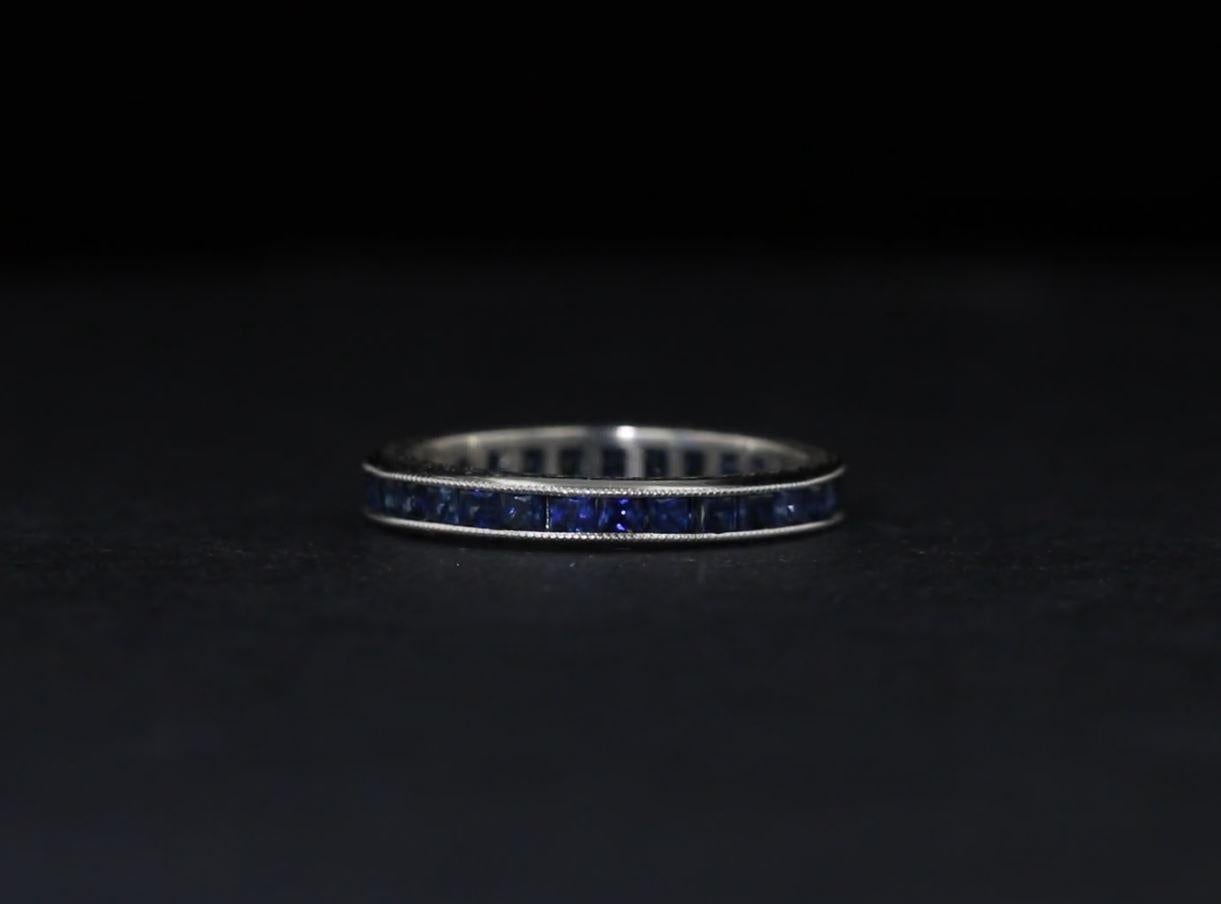 This estate channel-set eternity band is crafted platinum with engraved details along the ring's sides. The piece is set with approximately 29 square blue sapphires weighing approximately 1.50 carat together. The ring is currently sized to 6.