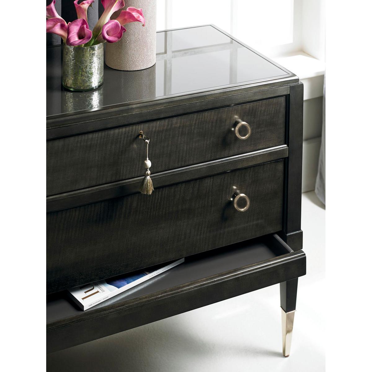 The clean, linear frame of this Charcoal colored nightstand gives off a streamlined air. The piece is punctuated by custom round hardware and tapered legs with gold ferrules. A dark glass top sits over three drawers, one of which is hidden for