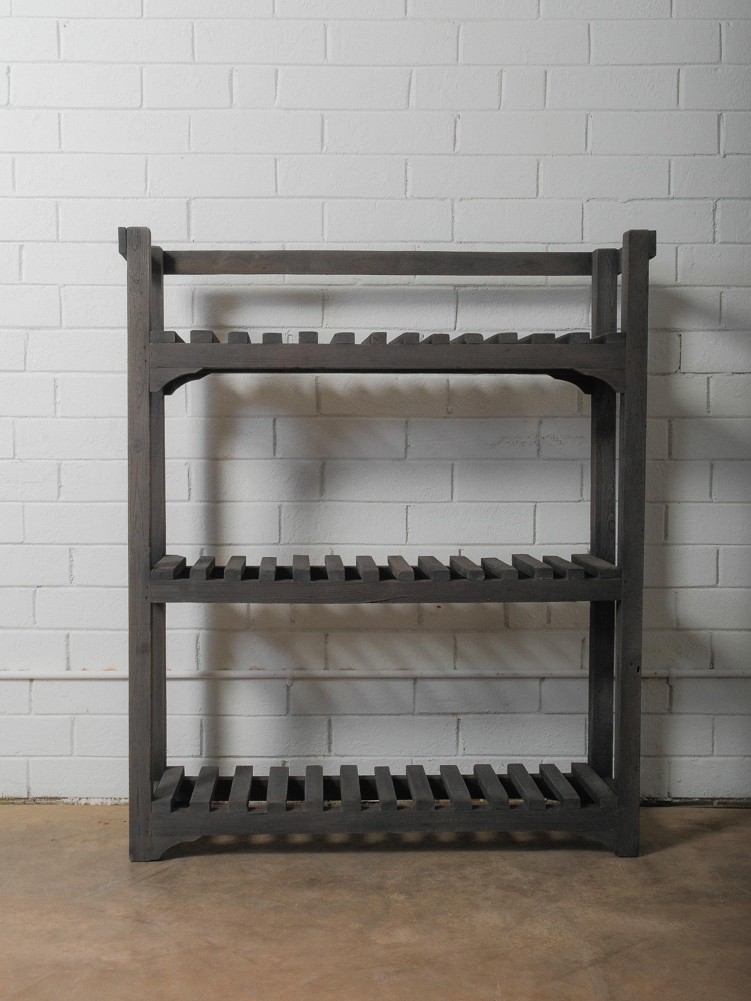 A linear study in repetition and form, this charcoal-toned wooden shelf presents a sculptural silhouette defined by its slatted construction. The open framework allows light to move through each tier, creating shifting patterns of shadow and