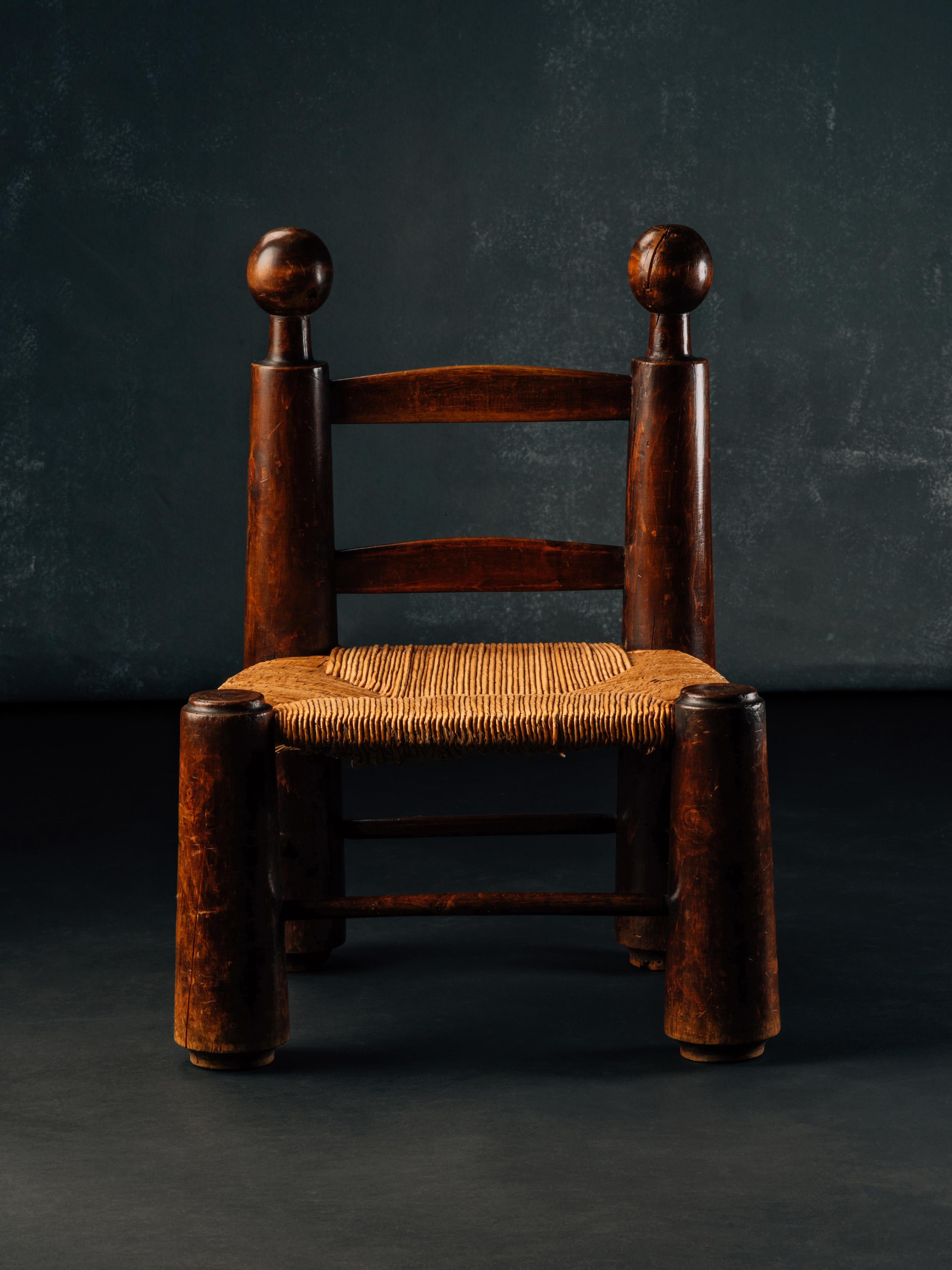 A low fireside chair (chauffeuse) or stool in stained oak attributed to Charles Duduouyt, with turned, truncated cone-shaped legs floating over recessed feet, turned stretchers, arched rails, and a woven rush seat. This example is a particularly