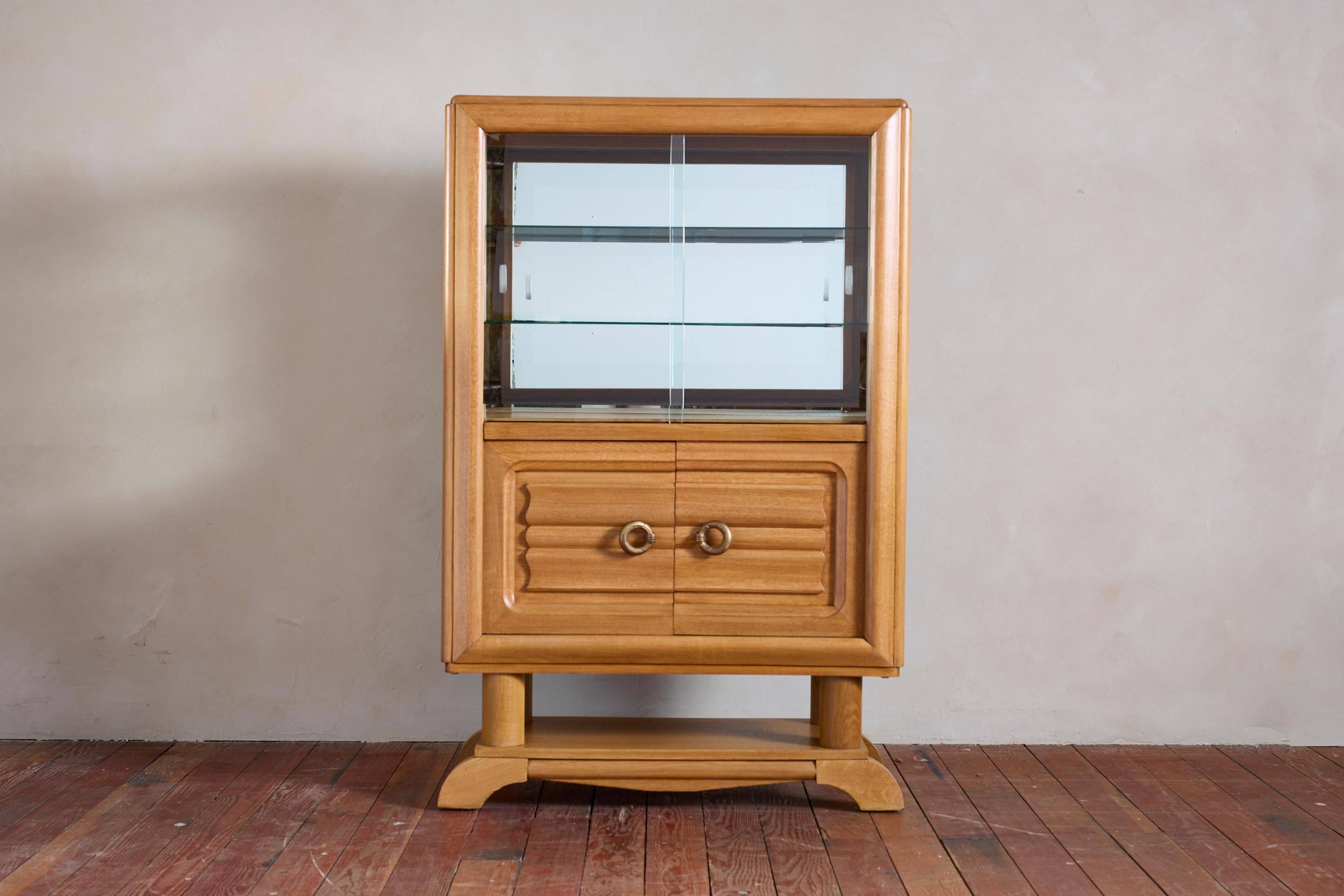 Charles Dudouyt style vitrine with glass upper sliding doors and interior mirror with 2 glass shelves. 

Lower doors are carved with signature Dudouyt style pattern and open to reveal interior shelving with inlay detail. 

Large cast brass circular