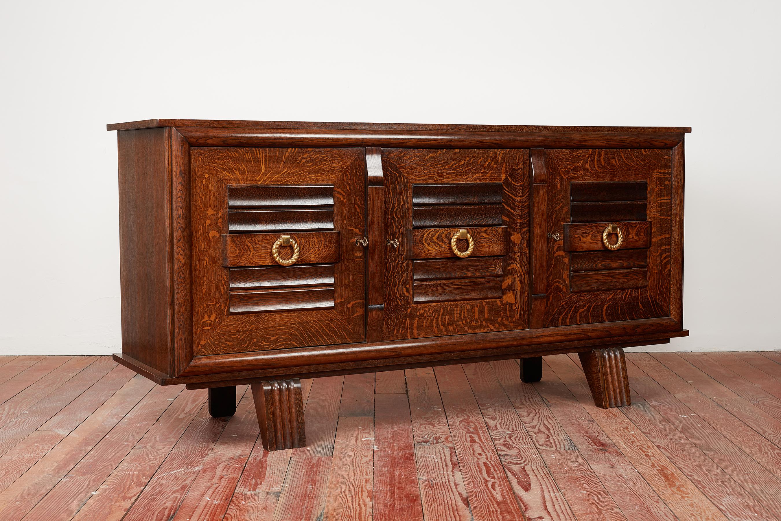 Charles Dudouyt sideboard in beautiful oak patina and carved linear detail 
Three cabinet doors with brass circle hardware open to shelving and a single drawer in center compartment area
Checkerboard inlayed top 
Floating on curved carved legs 
