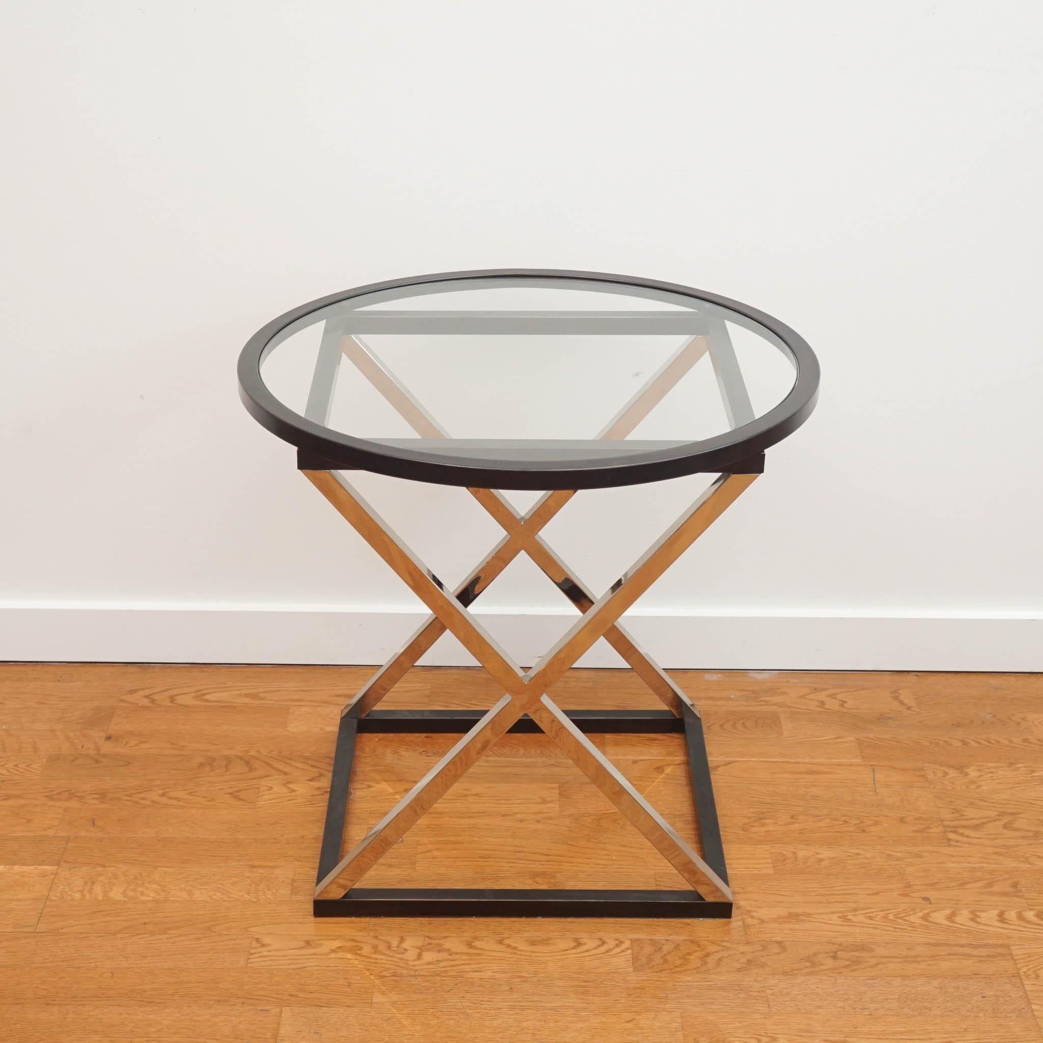 A perfectly proportioned metal side table with cross braced legs forming an ‘X’ shape and round top inlaid with clear glass, this design is both chic and versatile. 
This table is custom finished with black detail on base and around the glass
