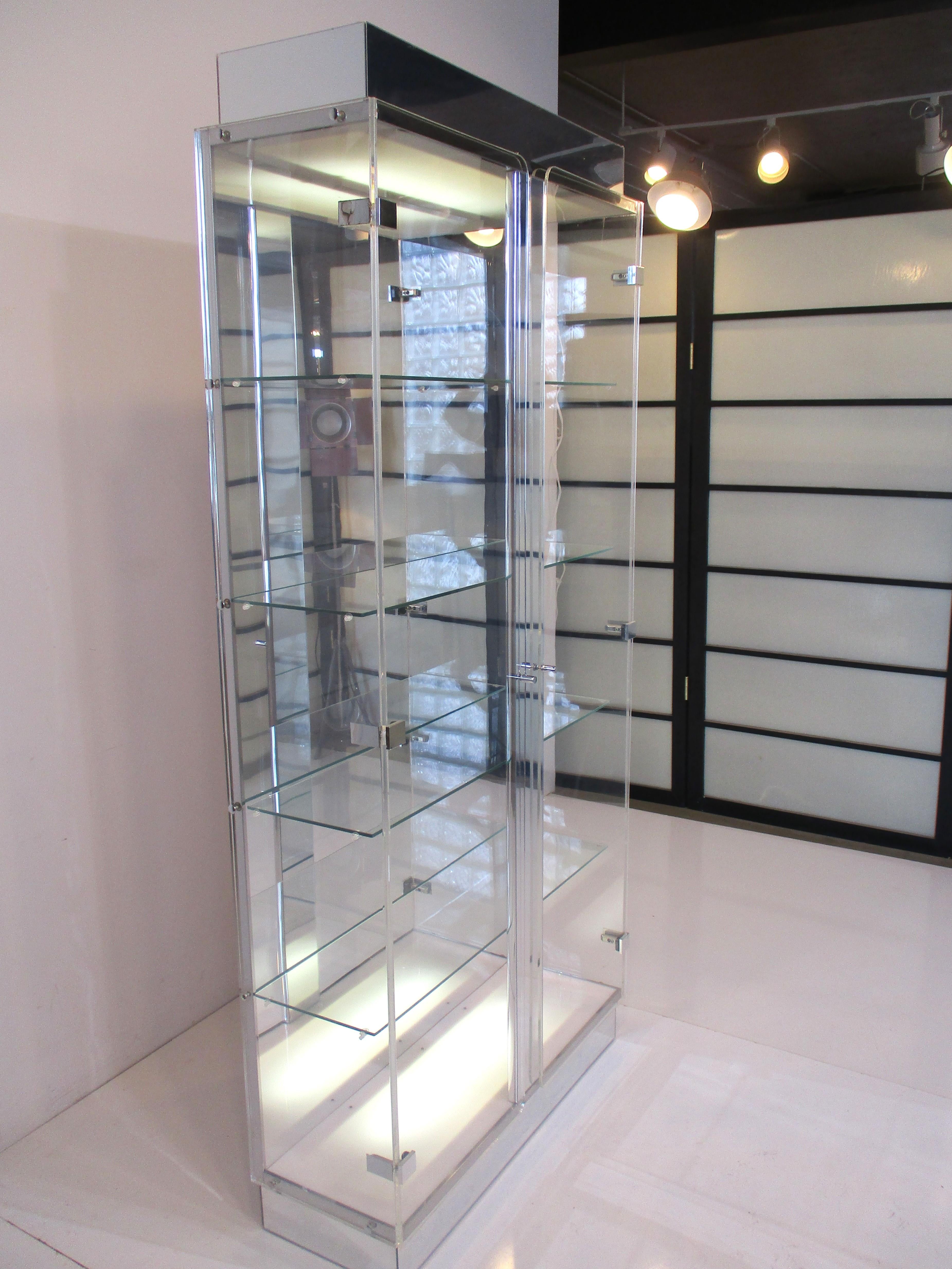 A two door Lucite display cabinet with lighted top and bottom panels having four glass shelves . The lower kick and upper top are in a chromed Lucite with chrome metal hinges and pulls . The cabinet has great presents with the down light and upper