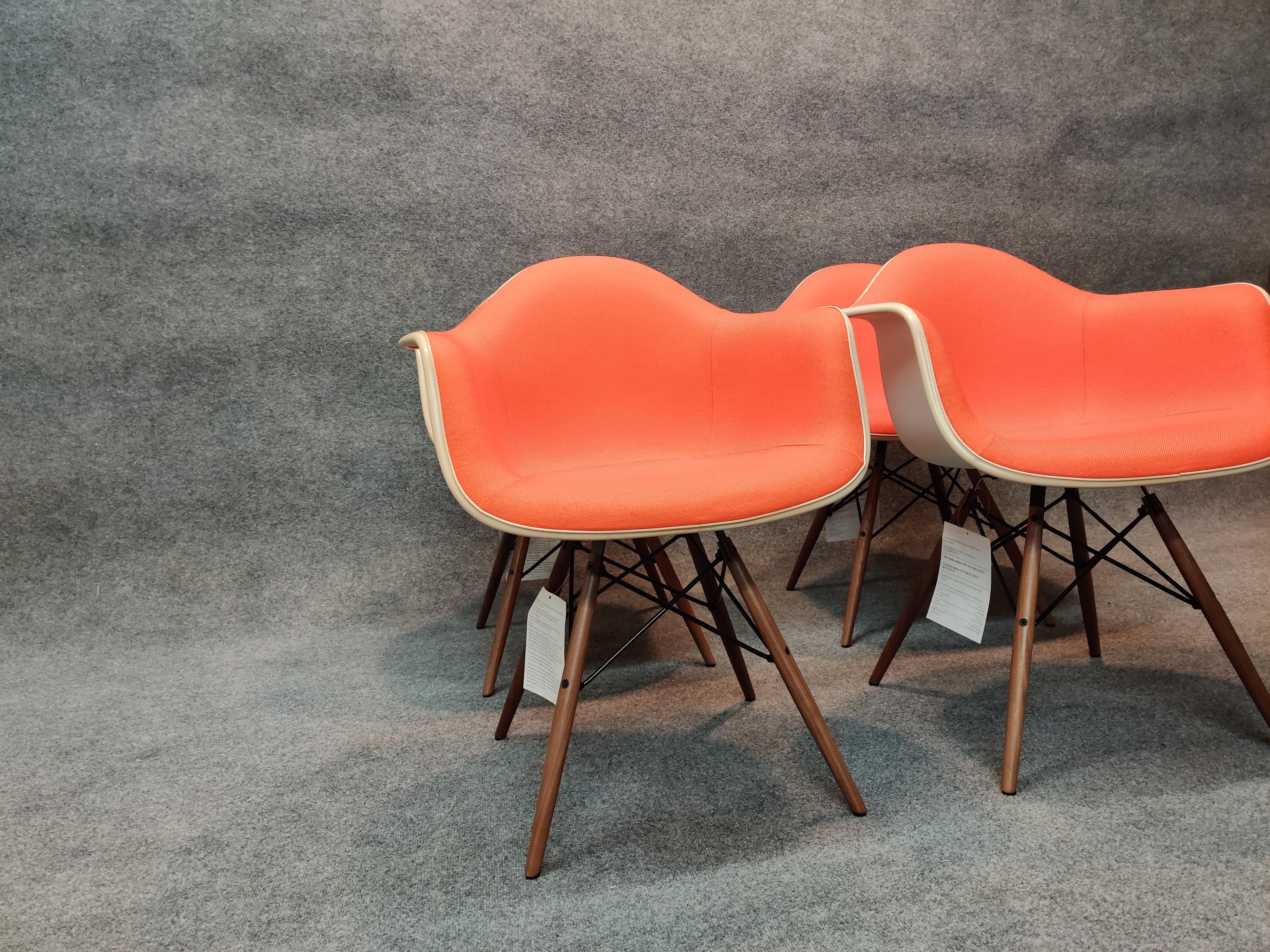 Designed by Eames, of recent manufacture by Herman Miller, these signed chairs are in like new condition. Each still retaining hang tags, shells are made of white plastic with an off-white band securing bright orange upholstery. Mounted on walnut