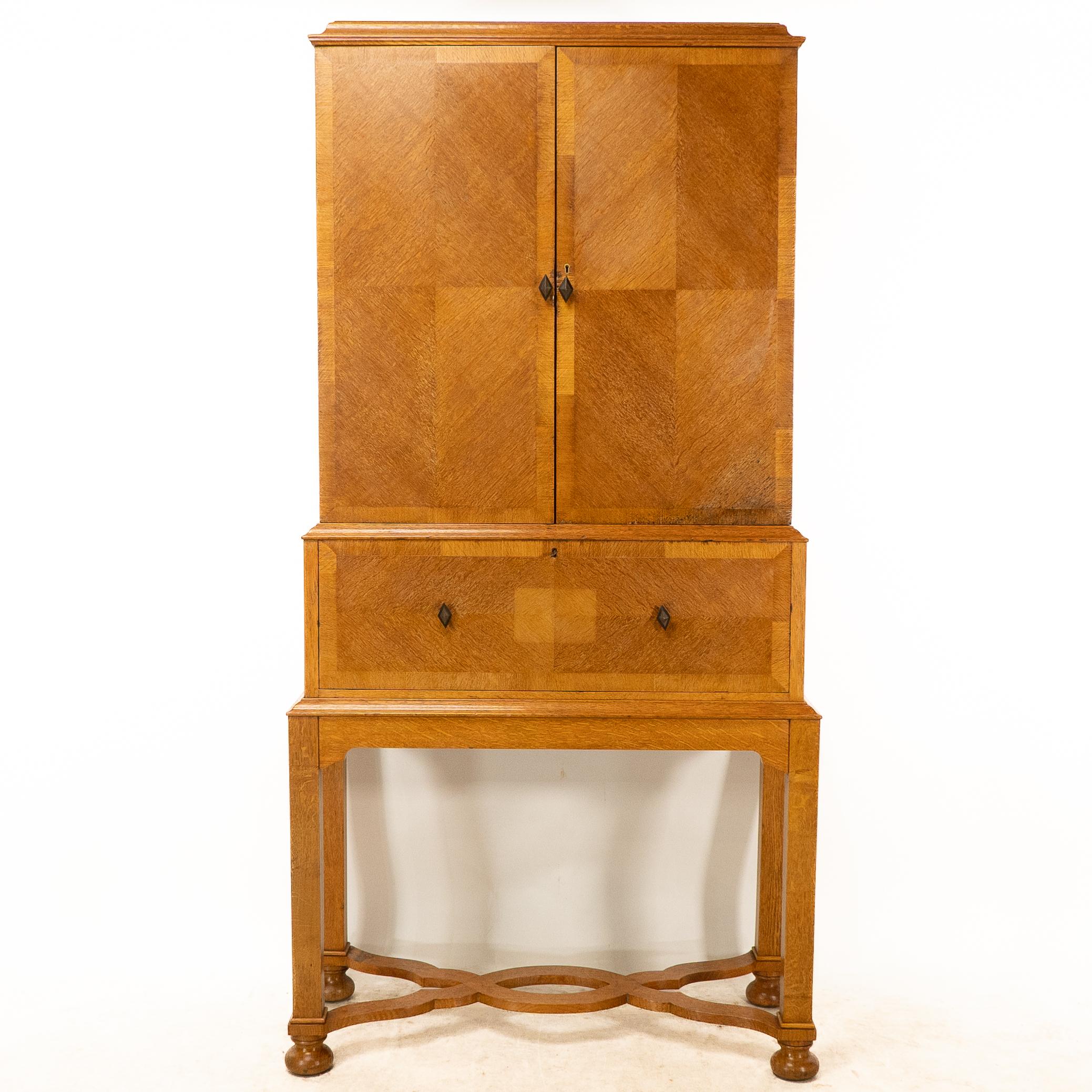 Charles Sydney Spooner, probably made by J H Brandt. 
A rare oak secretaire cabinet on stand, with quarter veneered panels to the doors and drawers with black and green diamond shaped handles. The doors open to reveal adjustable shelves. The sides