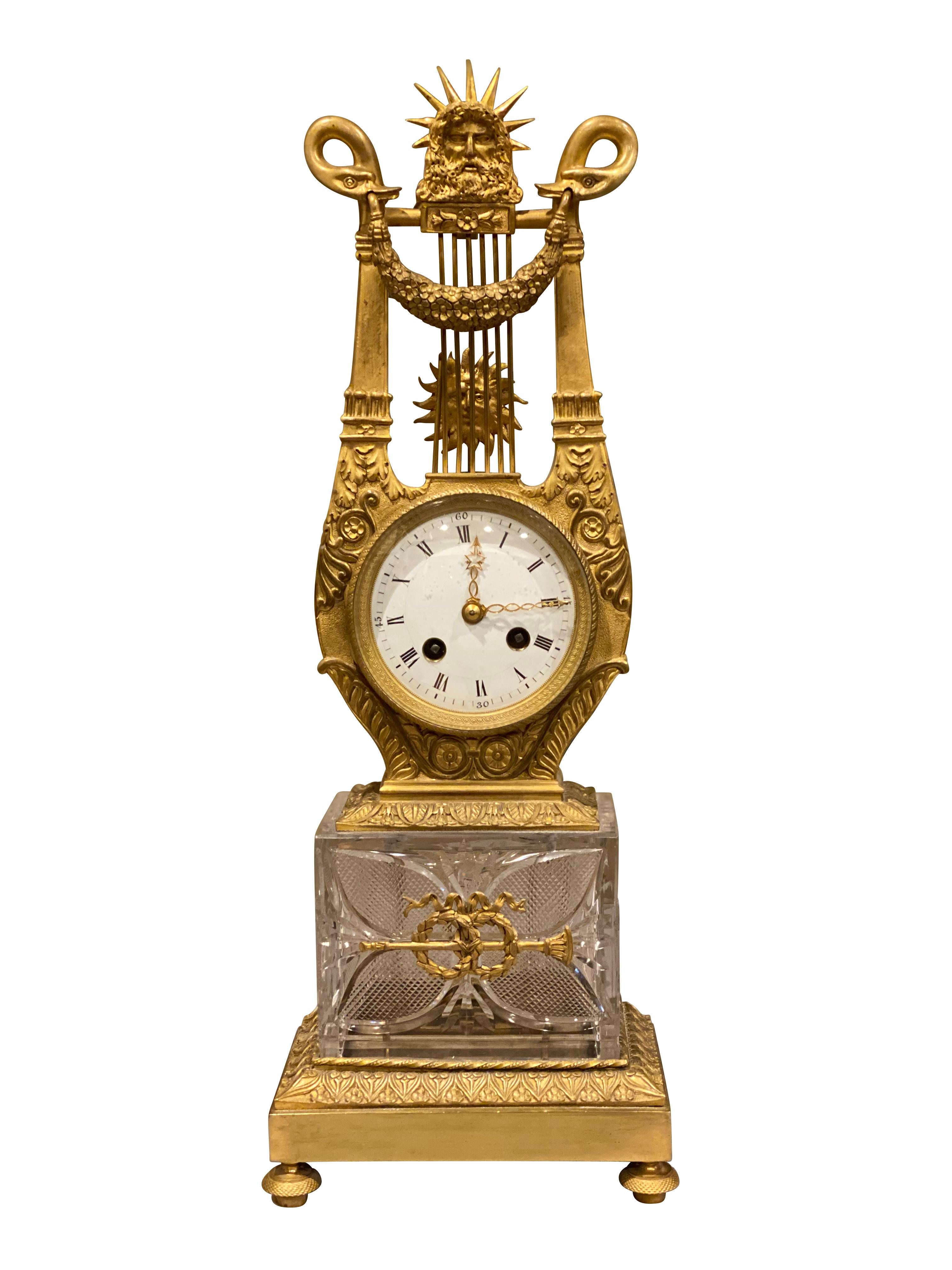 With sun god atop a lyre form case the lower part holding an enameled dial face above a cut glass and bronze base.