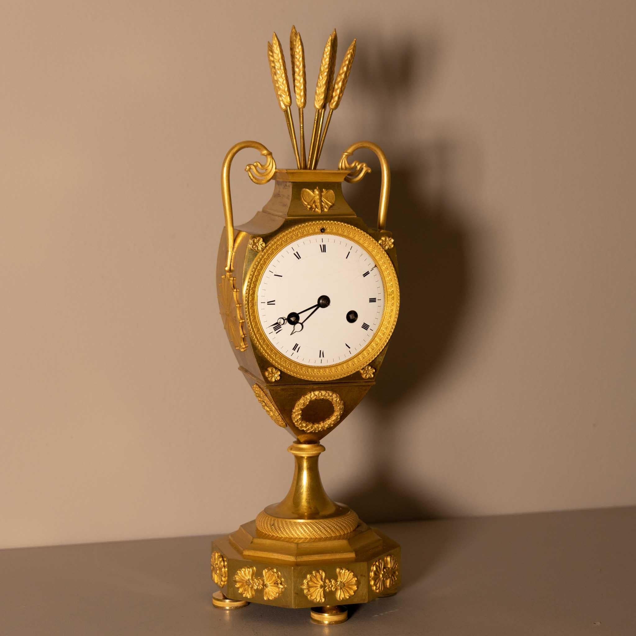 Fire-gilt bronze mantle clock with an amphora-shaped case on an octagonal stand and applied decorations in the form of ears of grain, velaria, laurel wreaths and butterflies. The enamel dial with Roman numerals.