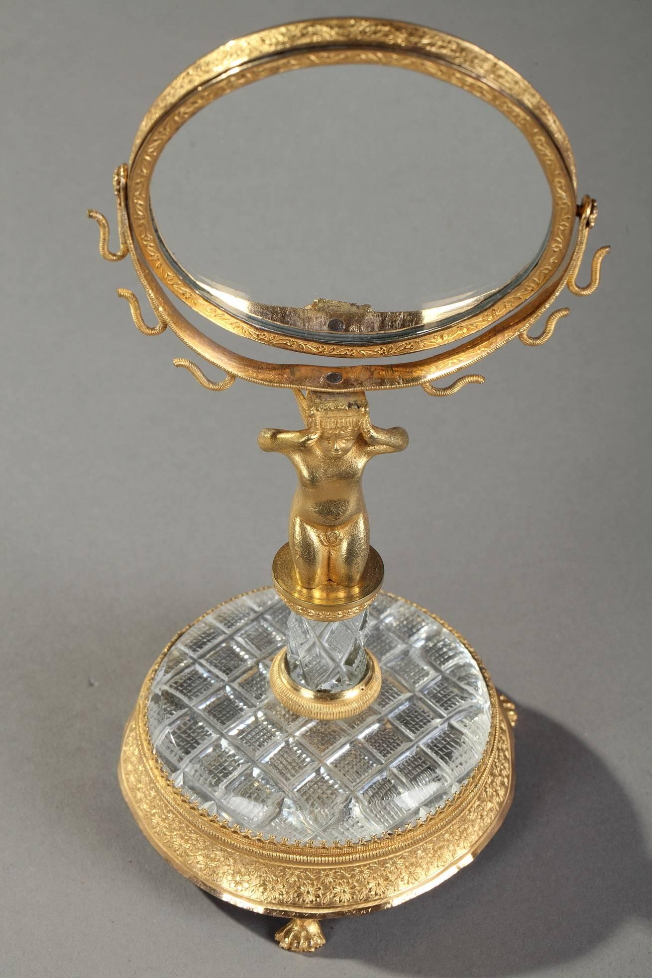 Gilt bronze mirror and ring holder depicting Cupid kneelig down on a small cut-crystal pedestal and base. He is holding the pivoting mirror above his head in his upraised arms. The mirror is adorned with small flowers and hooks to hold pieces of