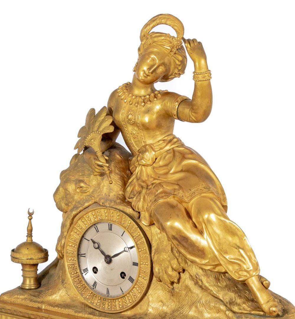 Charles X Gilt Bronze Figural Mantle Clock, circa 1830, the circular silvered dial, with strike on bell movement, surmounted by a reclining Turkish woman seated on a tiger skin and brazier beside her, on a rectangular acanthus leaf and scrolled base
