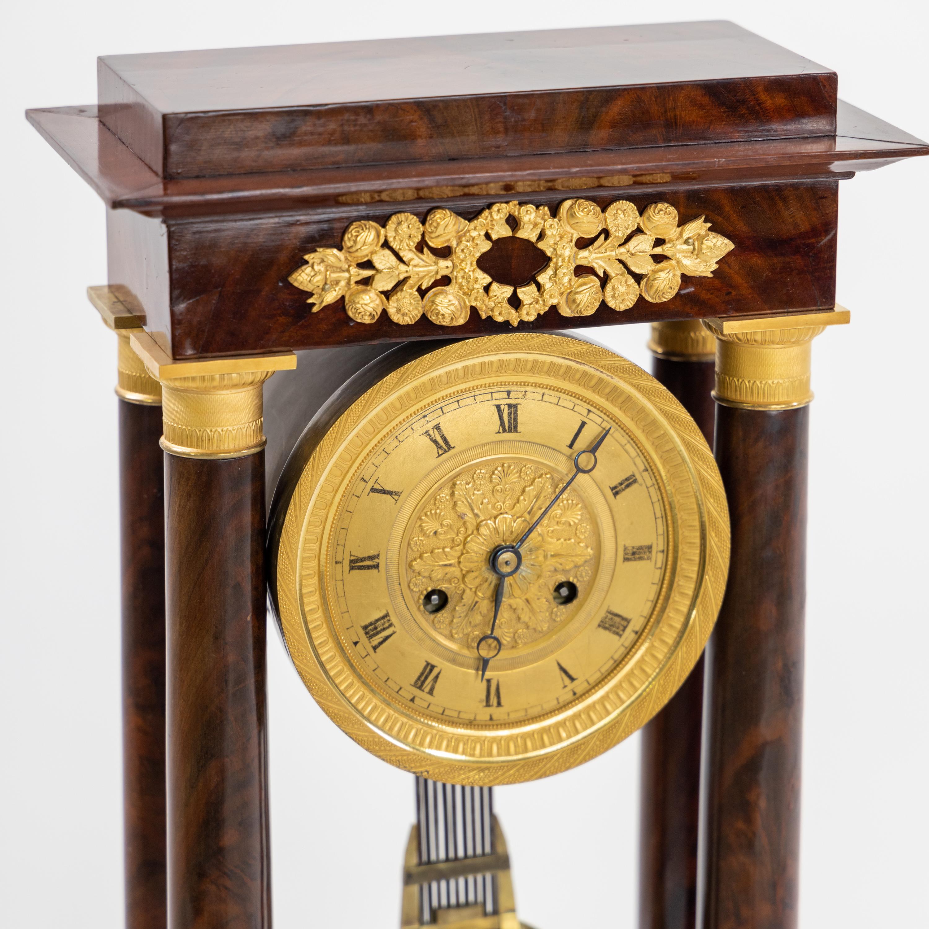 Portal clock on rectangular stepped base in mahogany veneer with small squeeze feet and fire-gilded applications in the form of rose festoons. The stepped architrave is supported by four smooth columns; the capitals and bases are made of fire-gilded