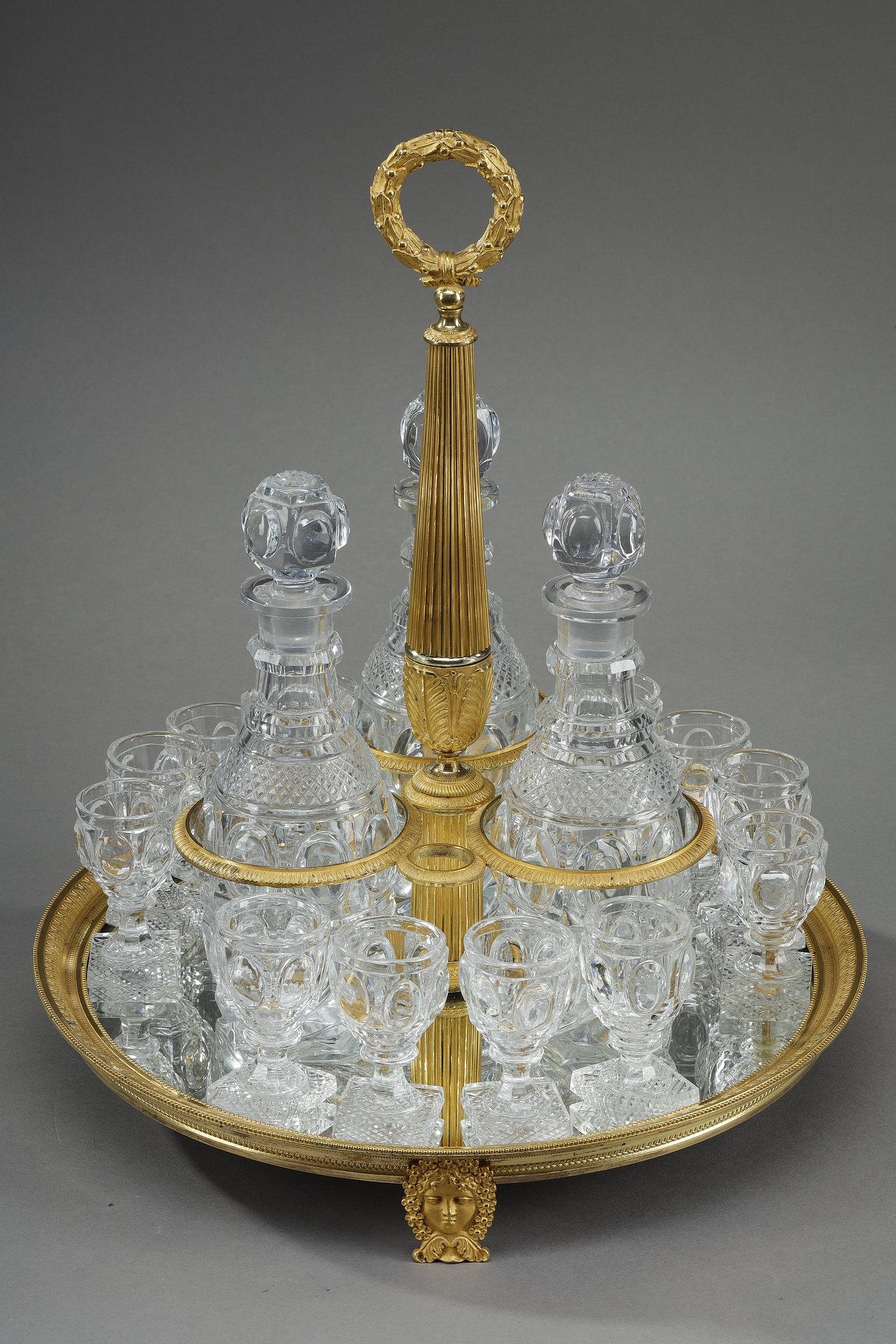 Cabaret en bronze doré à décor de frises de feuilles d'eau, de rangs de perles, la prise en forme de couronne de laurier surmontant un fût cannelé à palmettes et trois montures de bouteilles. Repose sur trois pieds masqués. Avec ses trois bouteilles