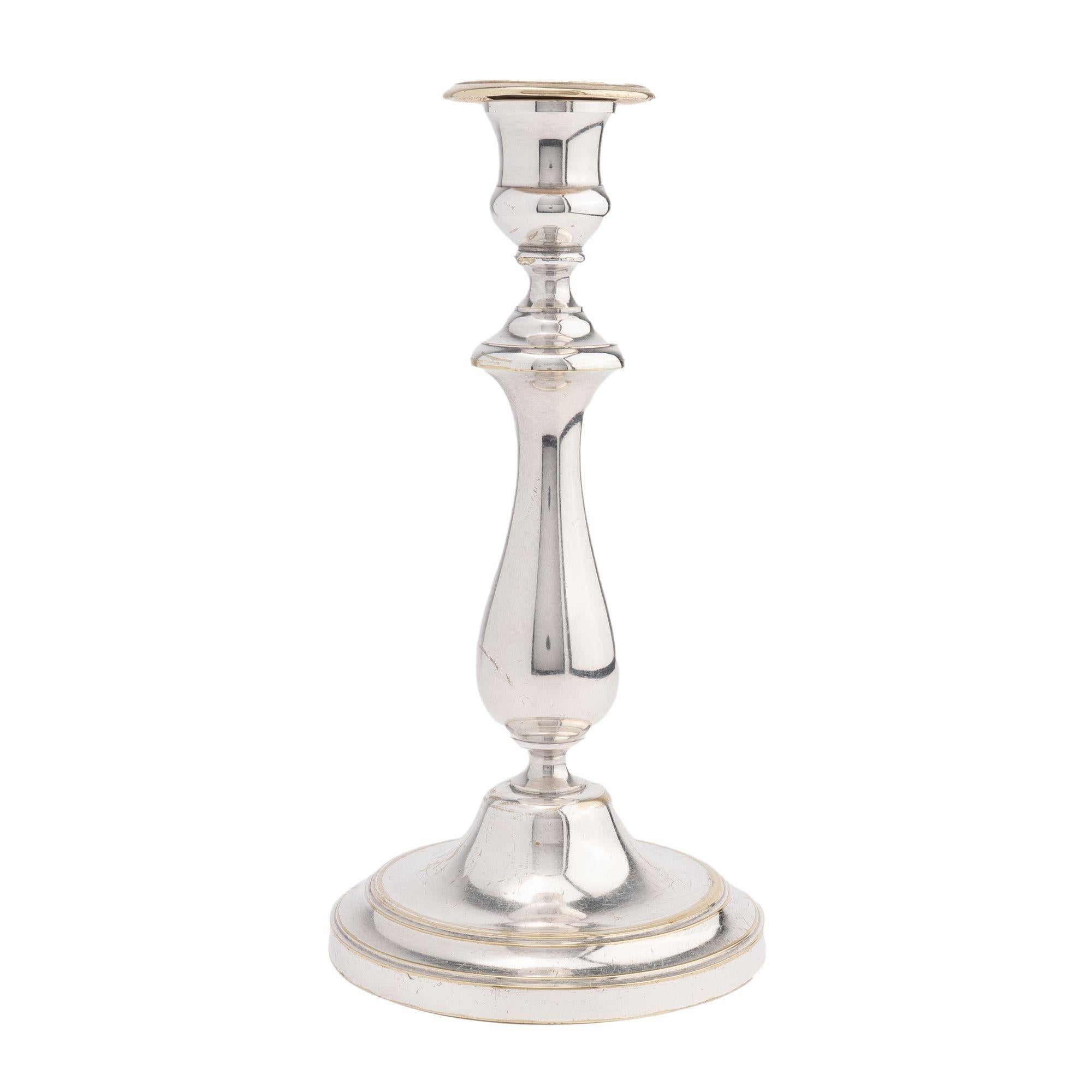 Charles X period silvered brass candlestick. The candlestick rests on a raised circular footed base, centering on a dome which features a waisted transition to the baluster form shaft. The shaft is topped by an urn form candle cup with bobeshe. The