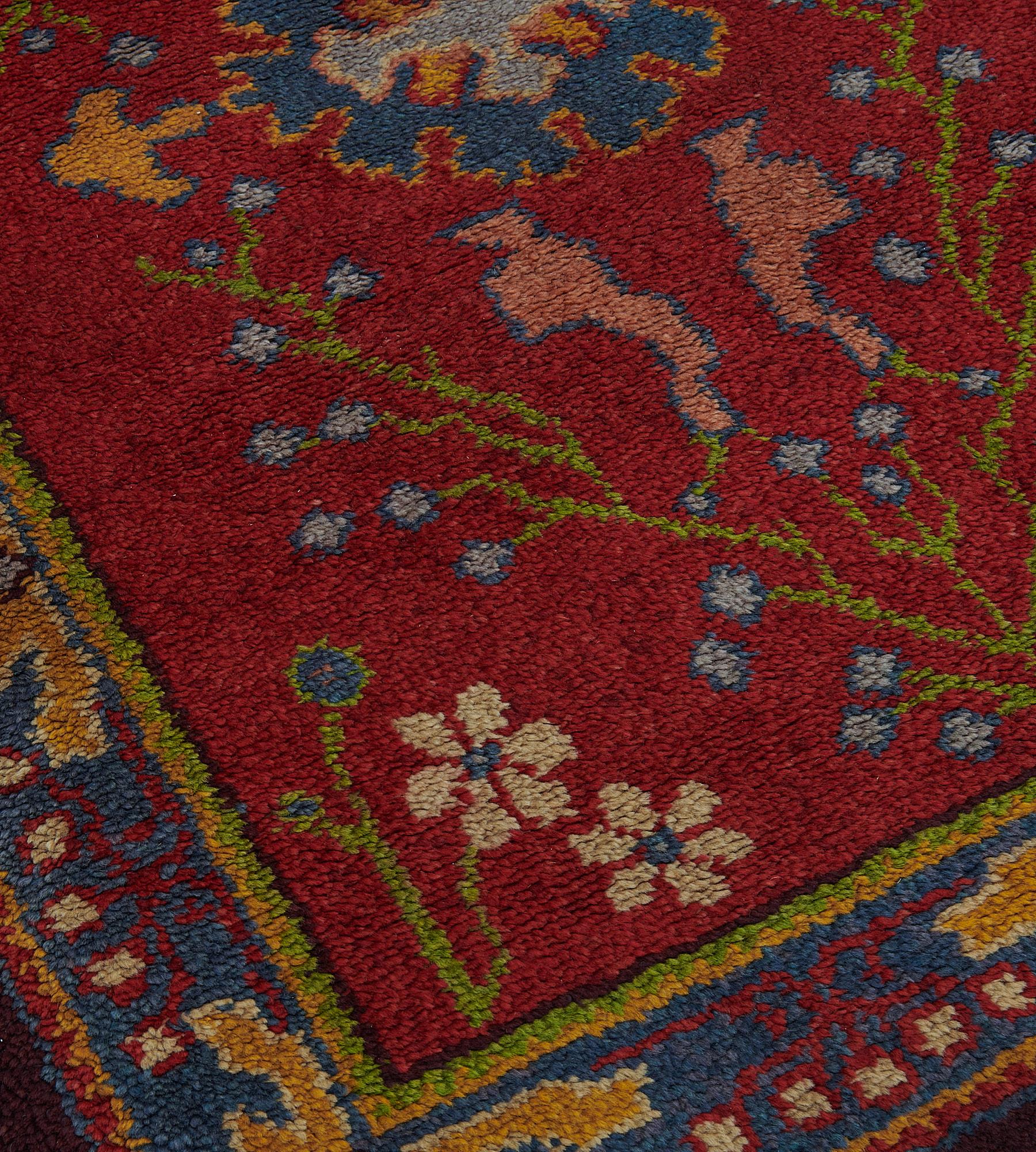 This antique, circa 1900, Irish Donegal rug has a deep red field with a central lozenge cartouche issuing at each side a mustard yellow part palmette with shaded pink flowerheads flanked by a delicate lime green tendril with daisy sprays and issuing