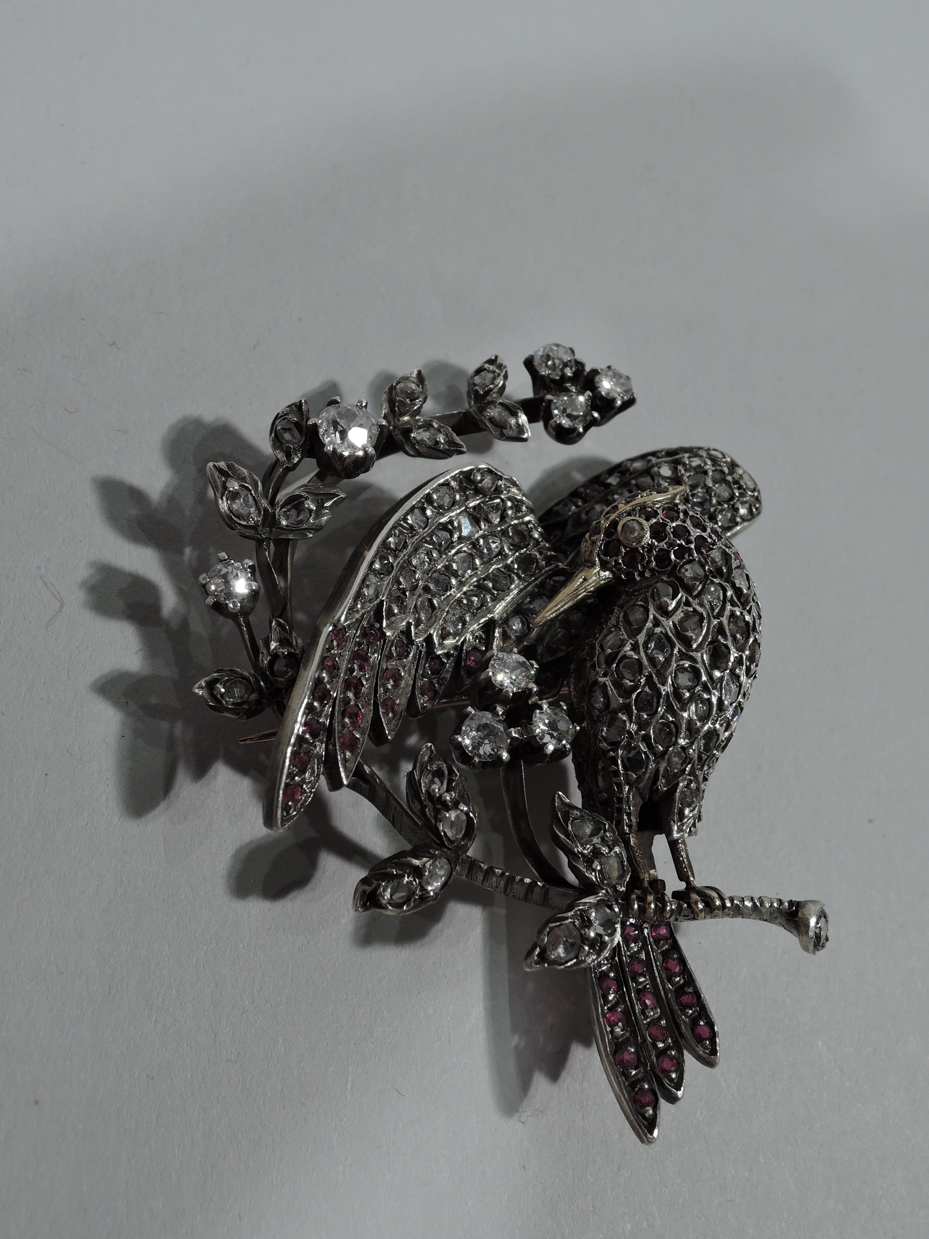 Charming figural silver brooch in form of bird. The bird is covered with inlaid rose-cut diamonds and faceted rubies on head, tail, and wing tips. Beak is gold as is ruby-encrusted crown – exquisite touches. The bird is perched on c-scroll branch