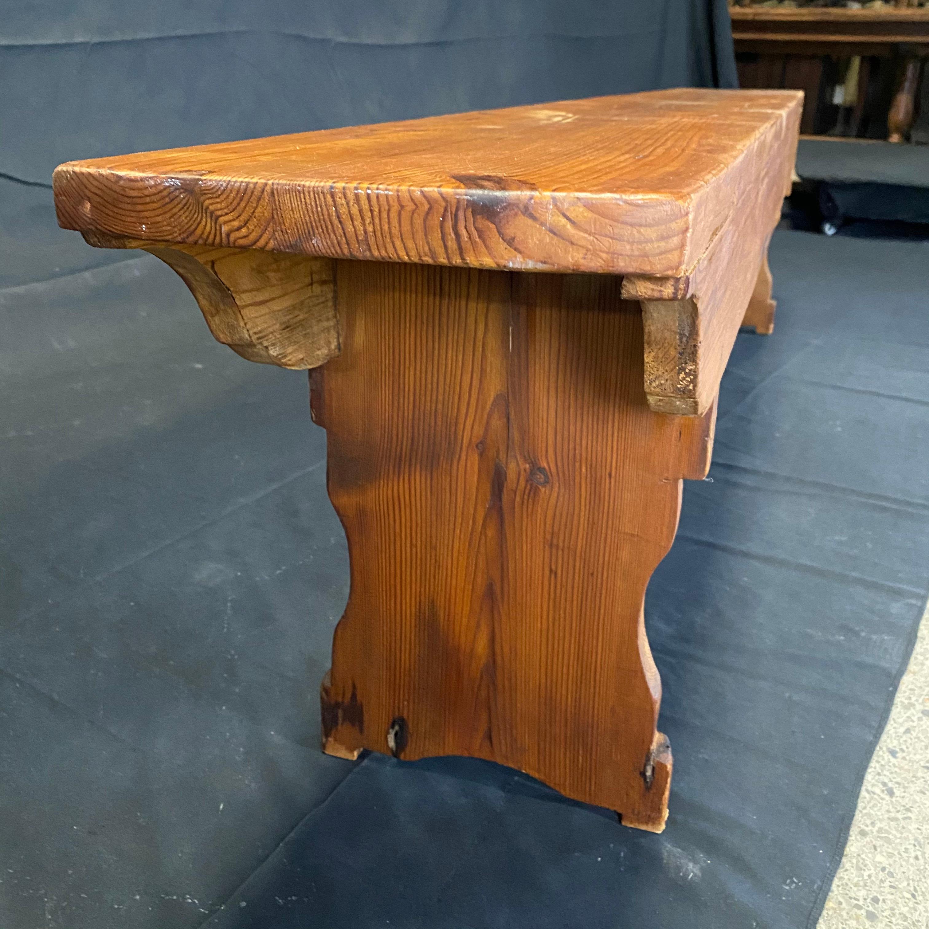 English long pine bench having a long rectangular seat which rests on two carved legs. The top of this bench is made from a single sturdy piece of pine, and on each side is a solid plank comprising the side aprons; the bench's solid construction