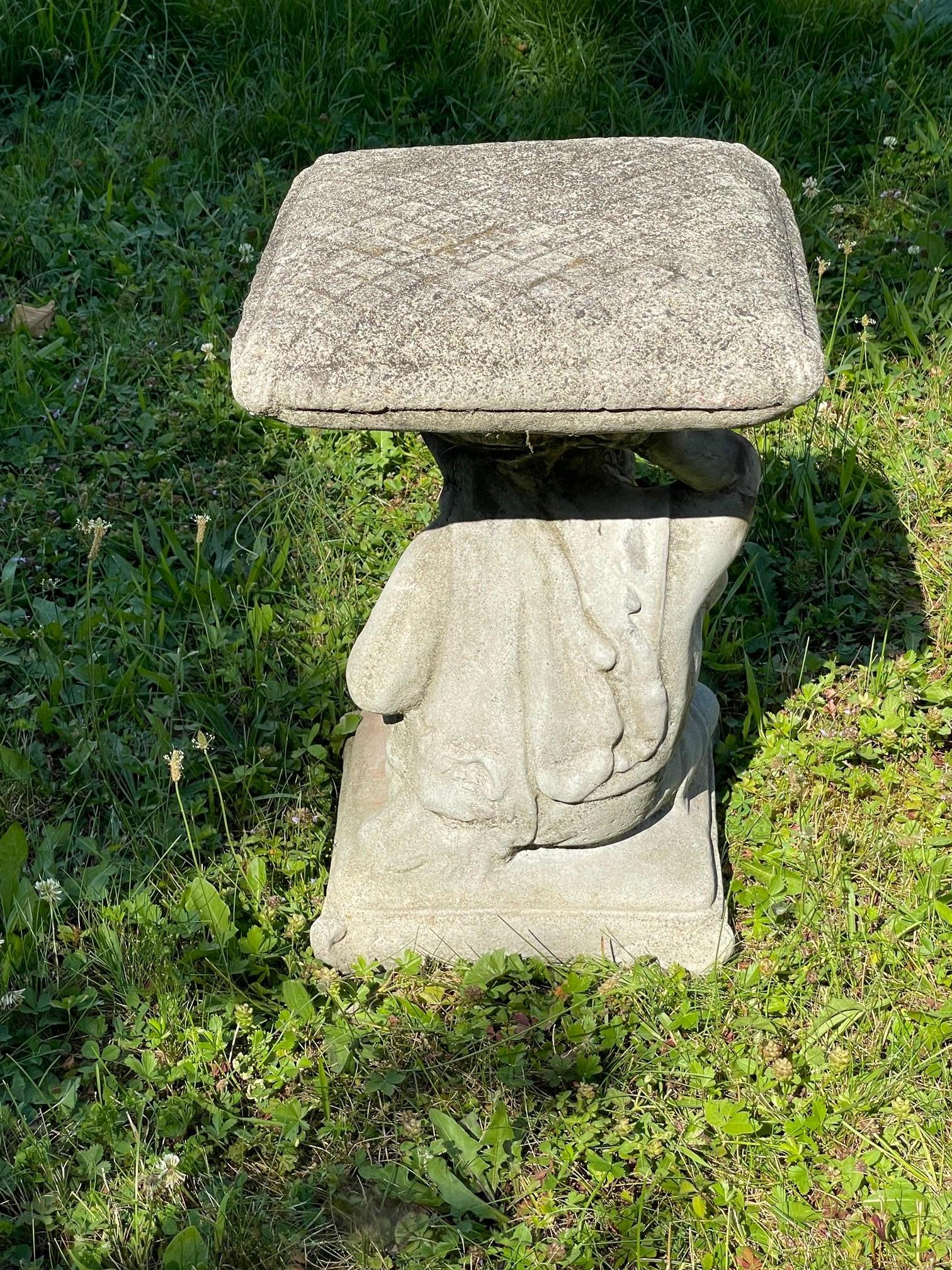 Ein wunderschöner Garten- oder Terrassentisch aus Betonguss mit einer sitzenden Putti als Sockel, die in charmanter Haltung mit einem angewinkelten Knie die kissenförmige Platte auf Kopf und Hand hält.