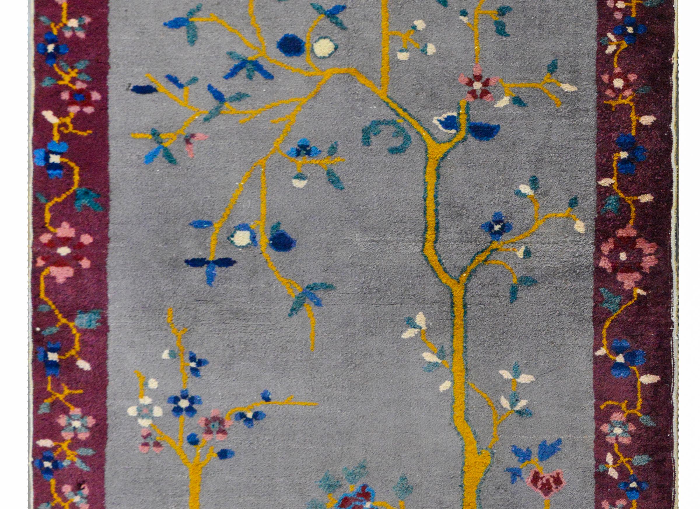 A charming Chinese Art Deco rug with a gray field surrounded by a violet border. The field depicts a flowering cherry tree perched above a scholars rock at the edge of a garden. The border contains a multi-colored floral pattern.