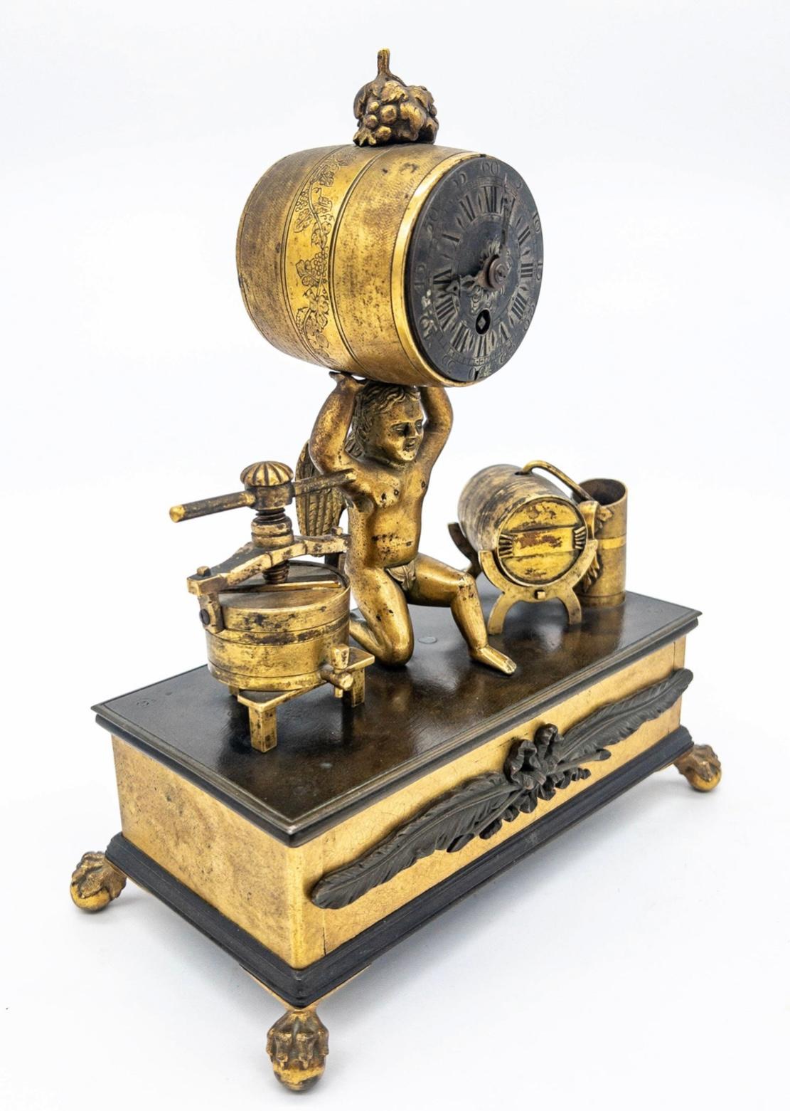A charming desk clock in the form of a cherub holding a barrel form clock with silver face, flanked by wine press and still, the base concealing a desk set with two glass bottles and silver caps.  The clock is overwound, but appears to be in good