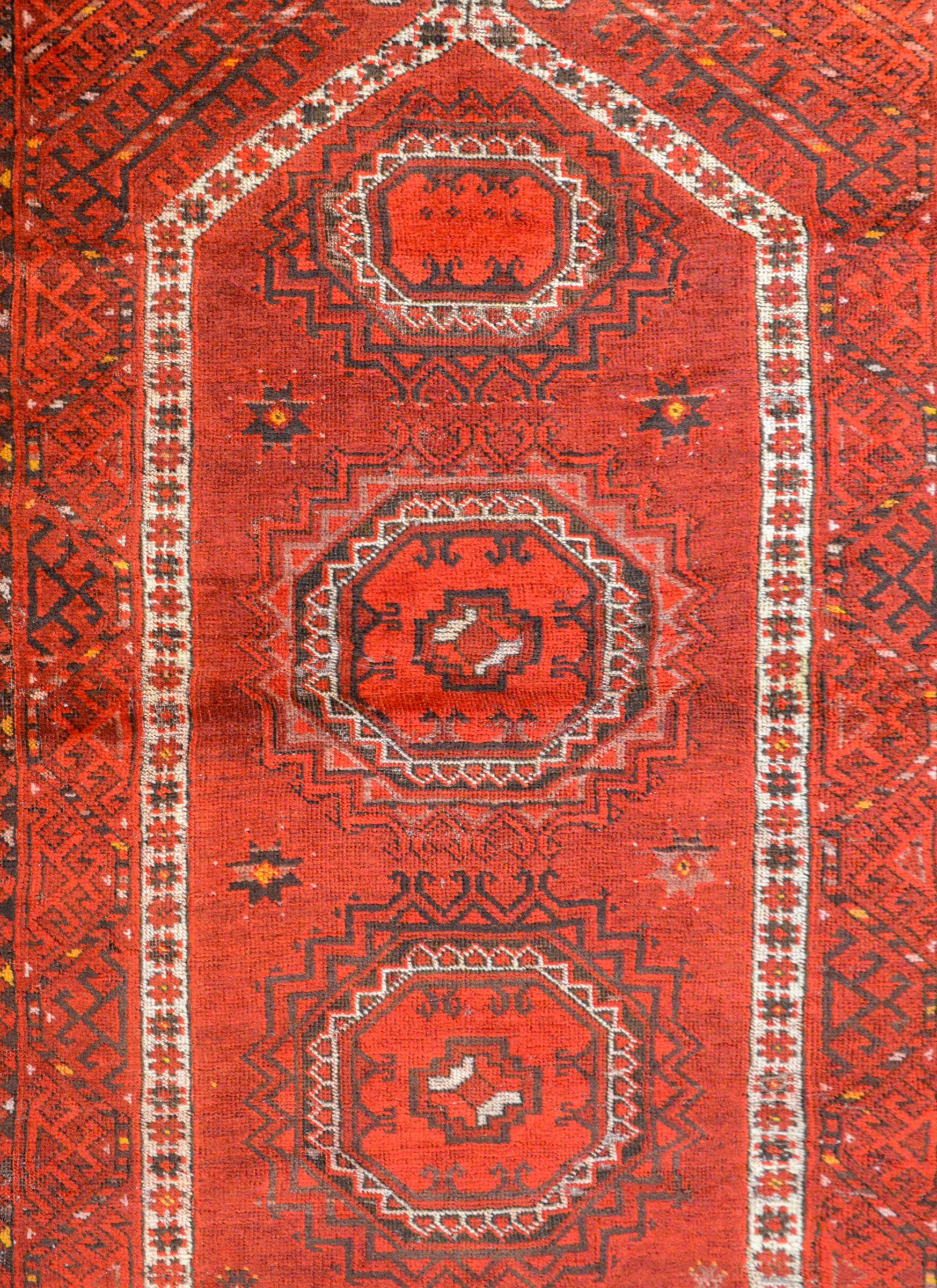 A charming early 20th century Afghan Ersari Prayer Rug with three octagonal medallions on a crimson background with geometric patterned halos amidst a field of stylized flowers. The border is wide with a fantastic tone on tone geometric pattern.