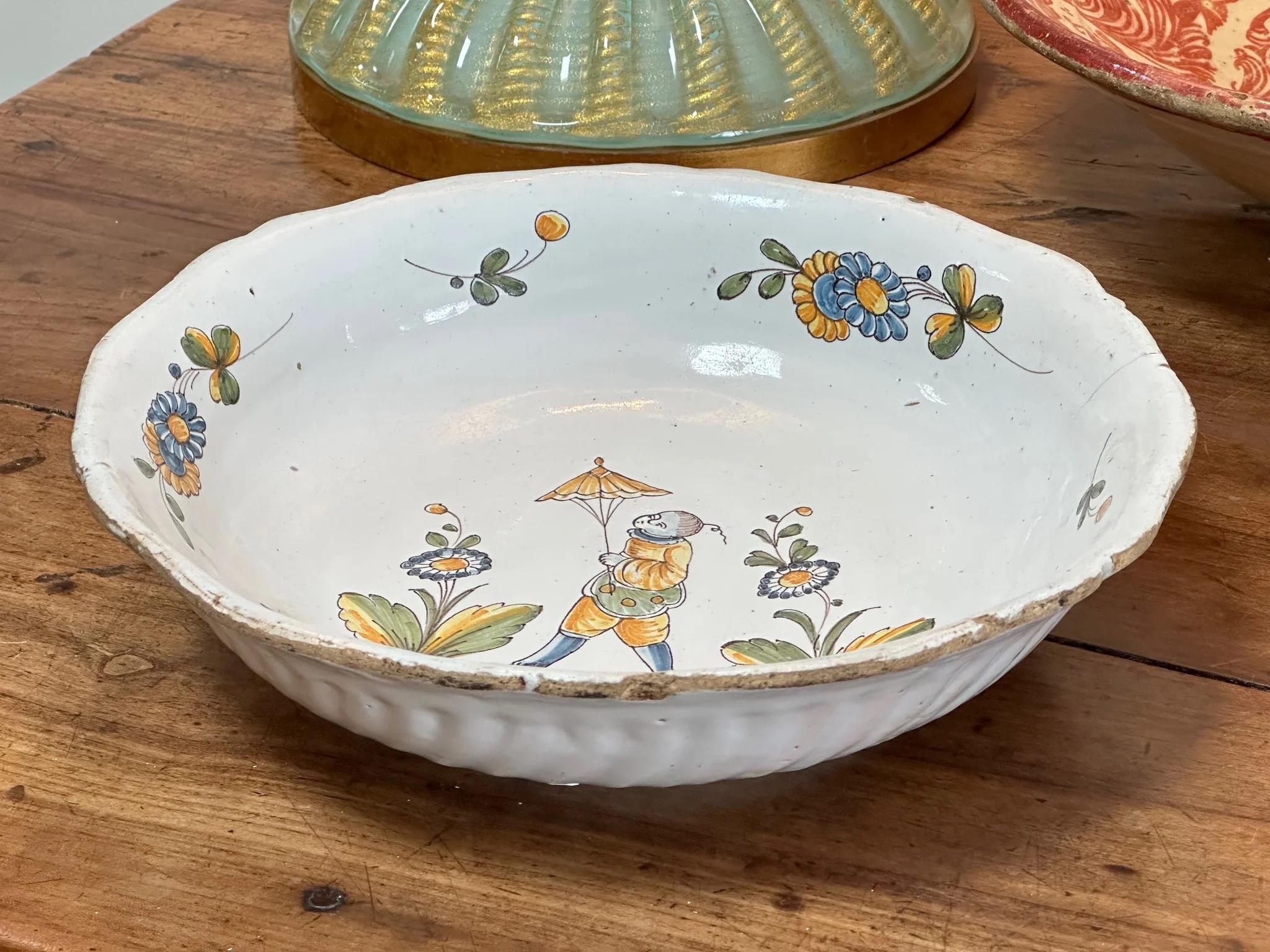 18th century Continental, most likely French,  Faience Earthenware Serving Bowl with fluted sides; hand painted vignette of a chinoiserie character with an umbrella flanked by images of flowers. Scalloped rim. Hanging holes on the foot.