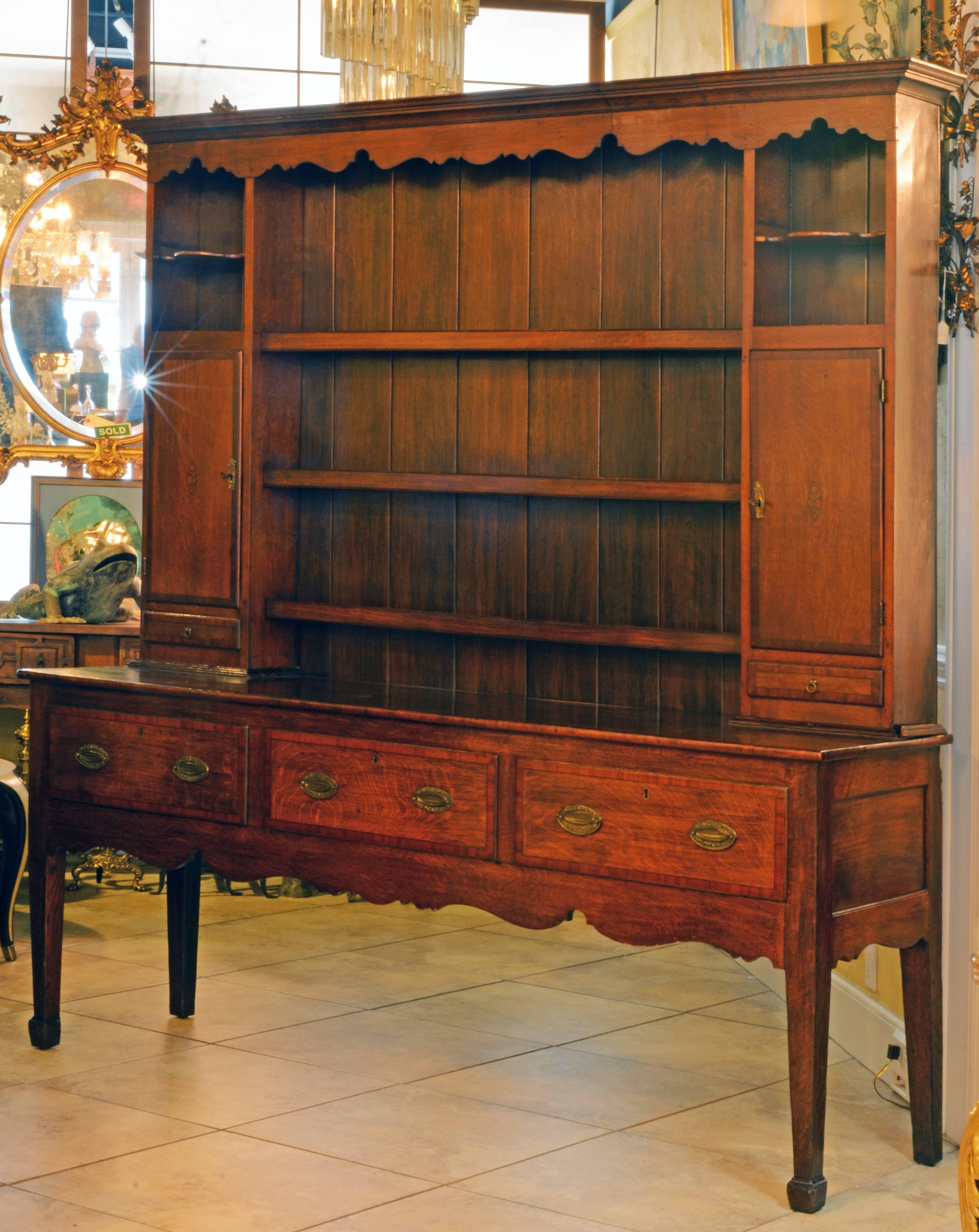 This welsh dresser consists of two parts. The upper part features a moulded corniche and a scalloped frieze above three plate rack shelves flanked by cupboards and small drawers with mahogany banded fronts above which smaller open compartments with