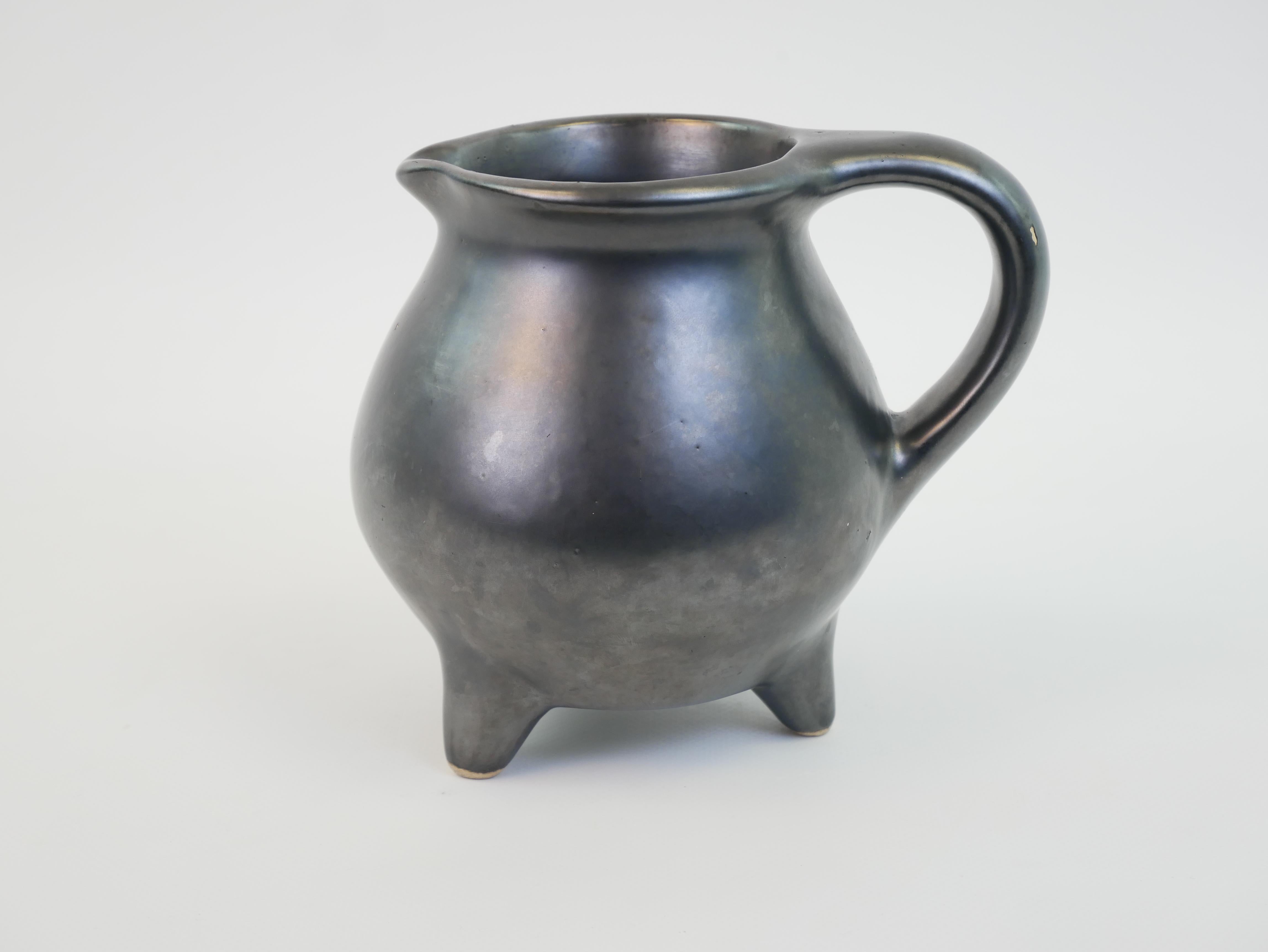 Esta pequeña y elegante jarra, que recuerda la obra de Georges Jouve, presenta una forma refinada y curvilínea con un lustroso esmalte metálico negro. Mide 12 cm de diámetro, 14 cm de altura y 16 cm de longitud incluida el asa, y se asienta con