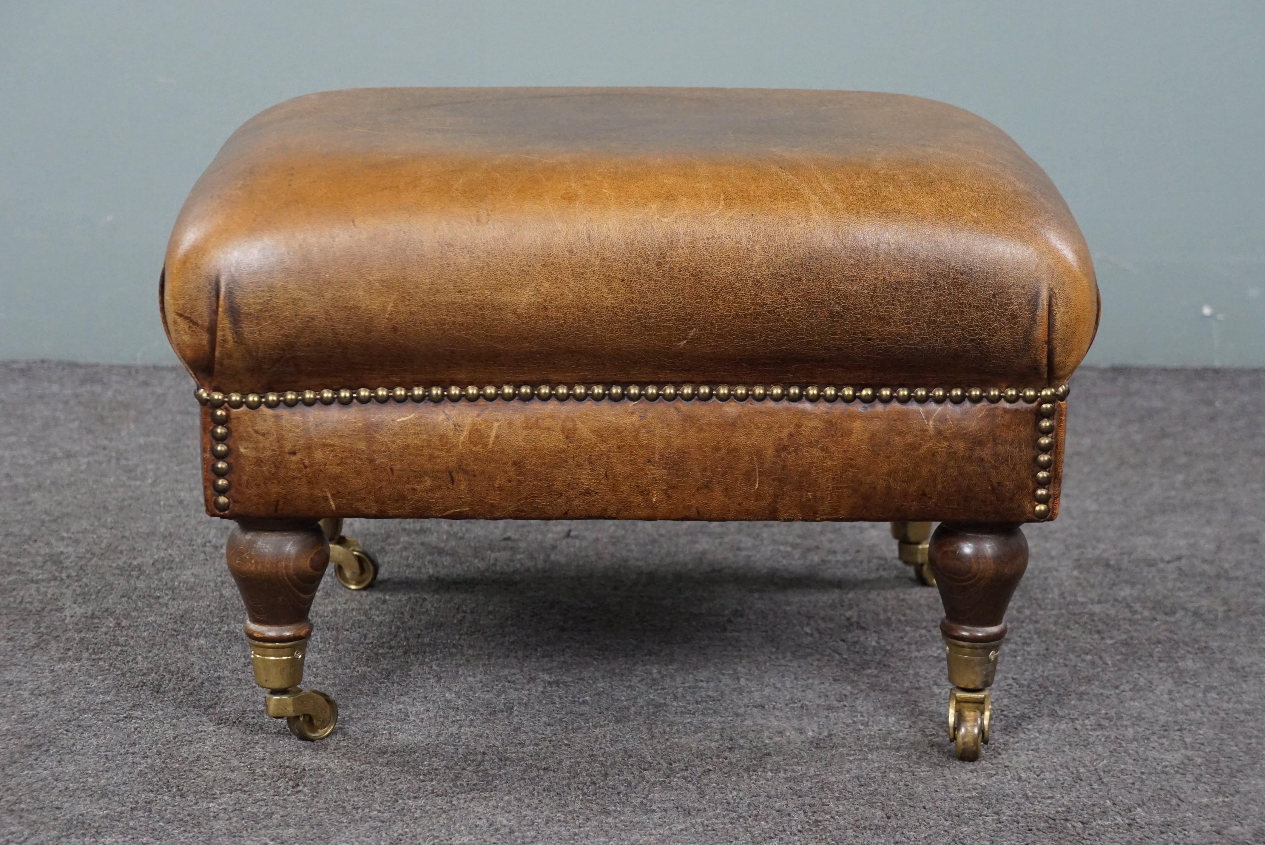 Offered is this charming sheep leather ottoman features elegant legs with brass wheels. A sheep leather footstool, also known as an ottoman, turns your already comfortable armchair into a true relaxation paradise. Resting your feet on this ottoman