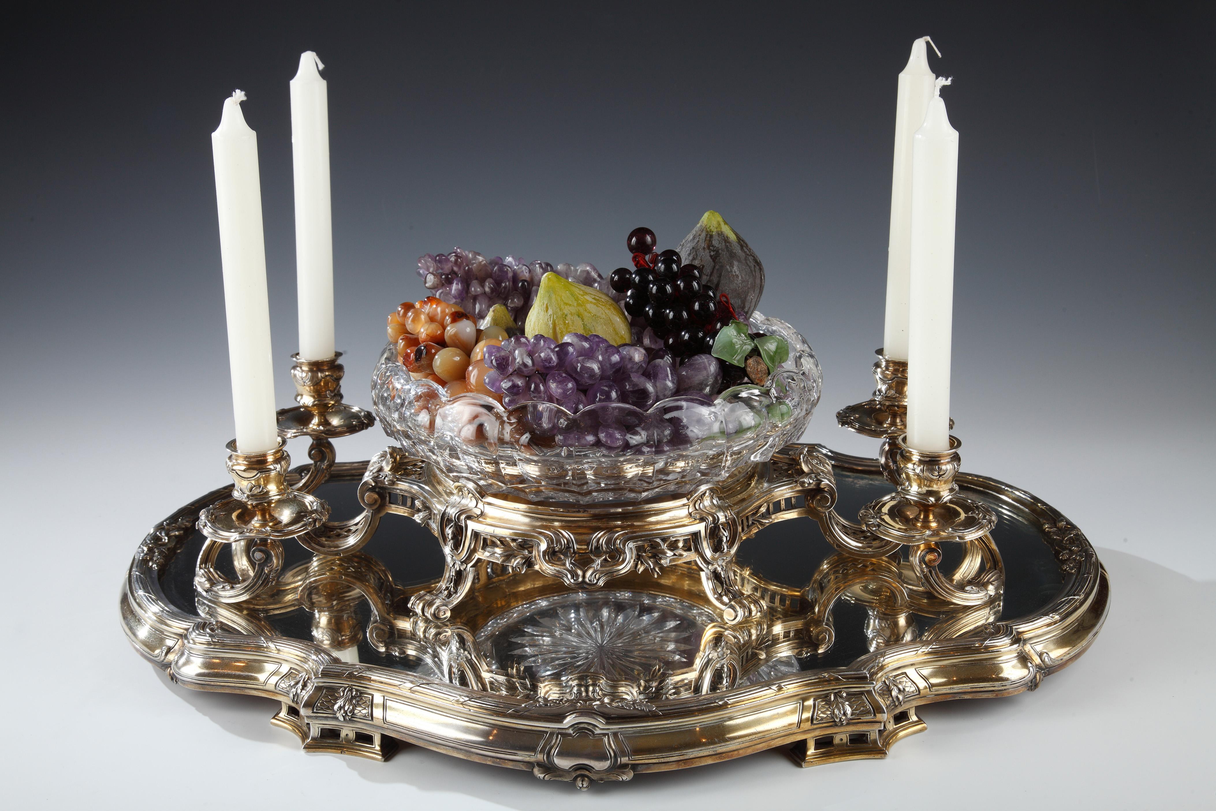 Elegante centrotavola in vermeil finemente cesellato. Composto da una coppa di cristallo tagliato Baccarat da cui fuoriescono quattro sinuosi bracci luminosi. Il tutto poggia su un vassoio smerlato a specchio ornato di fogliame, con un contorno