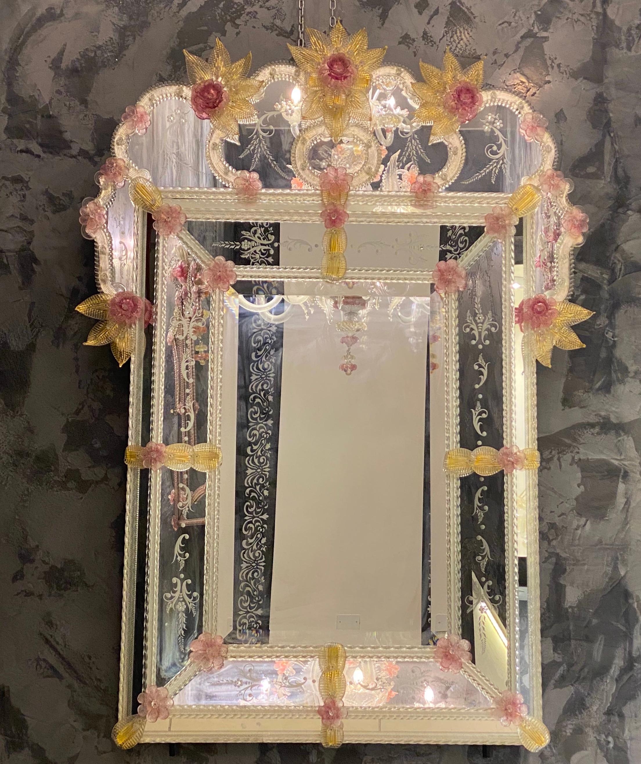 Ce charmant miroir vénitien présente des motifs floraux gravés qui ornent le cadre réfléchissant. Le long des bords du cadre se trouvent des accents de corde en verre et de nombreuses fleurs roses en verre et des feuilles d'or. 
Exécuté par le
