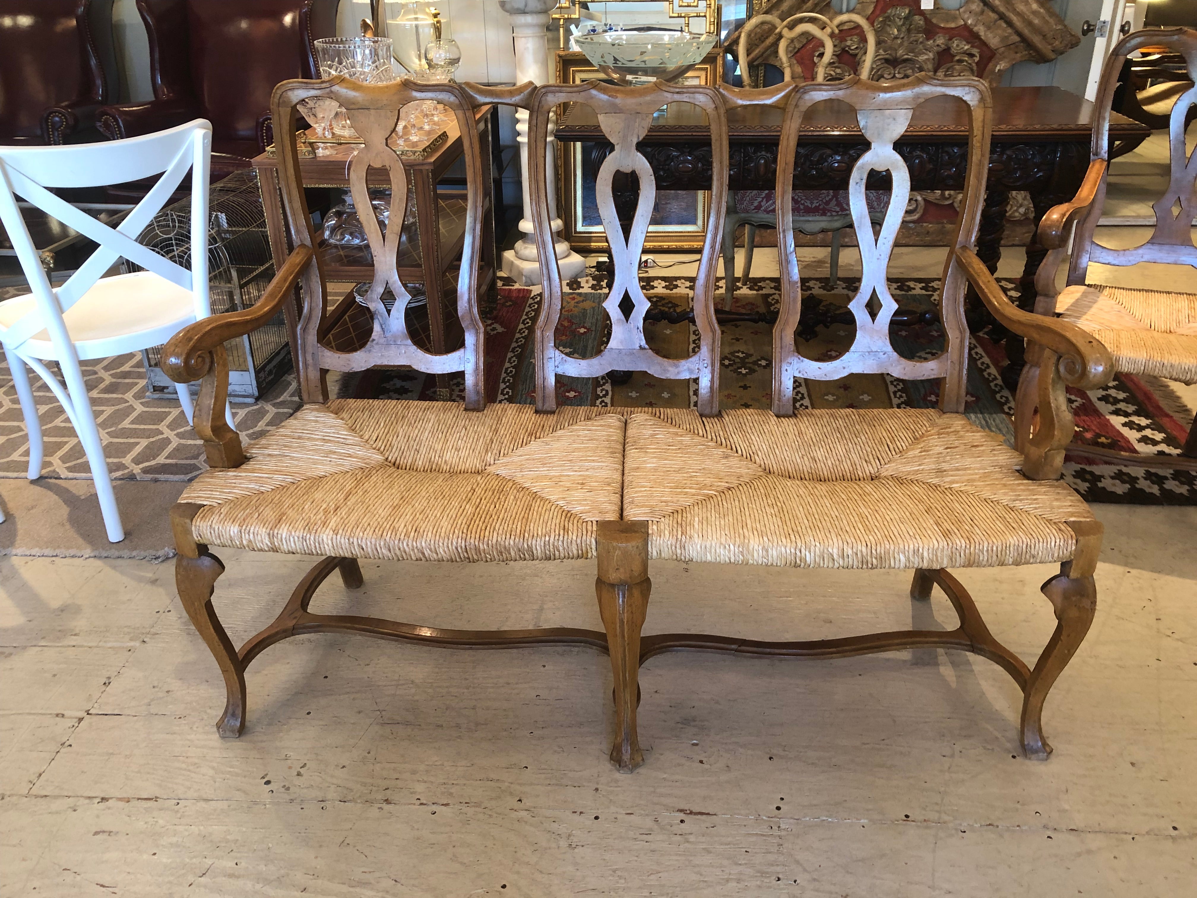 Large super charming vintage chestnut bench having scroll arms and 3 beautiful carved wood sections of the back. Seat is handsome original rush in great shape. Cabriole legs and stretcher underneath finish the French Provincial look with